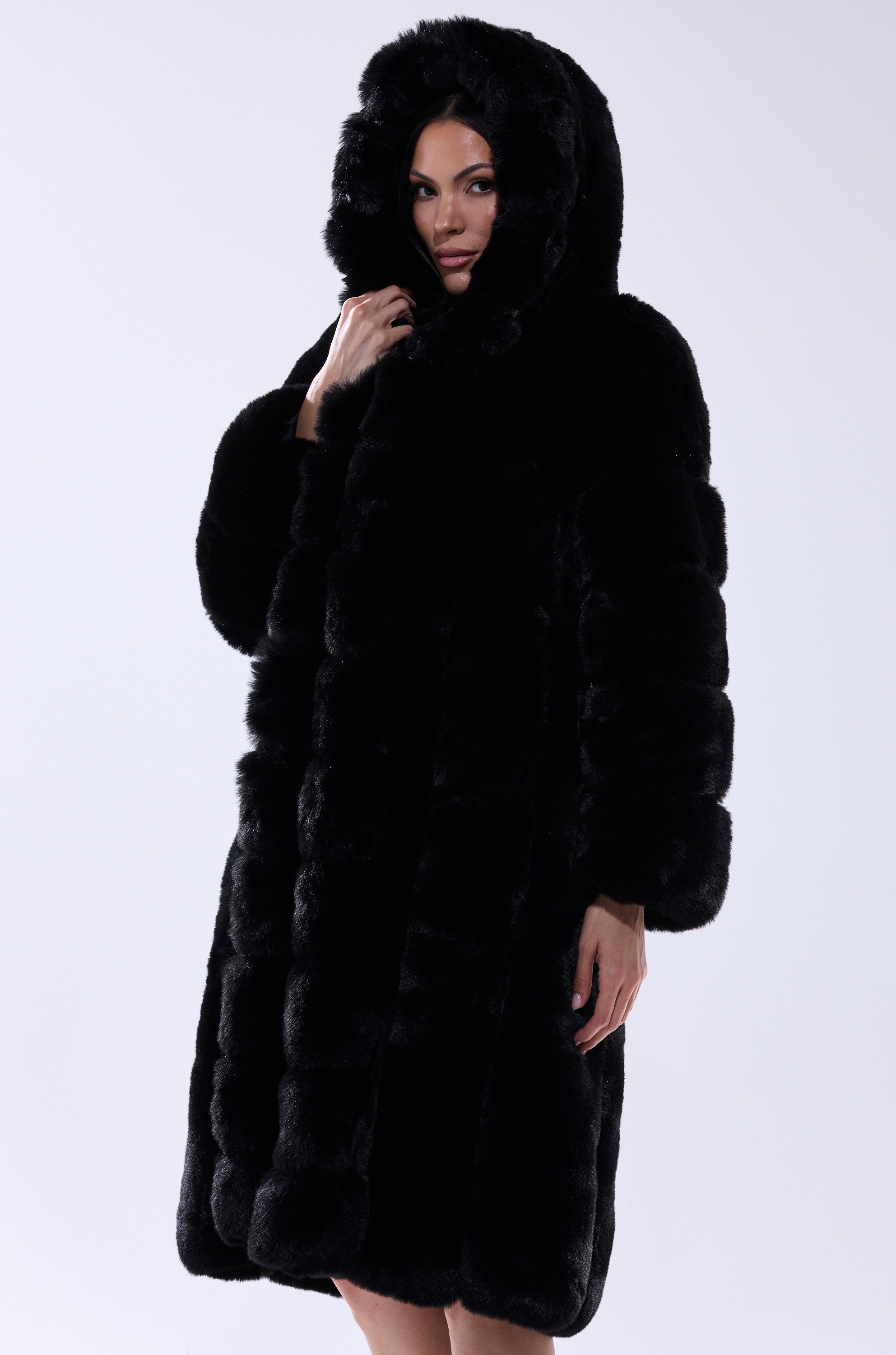 Wearing the BERLIN HOODED FAUX FUR IN BLACK, a person stands against a plain light background, partially lifting the large hood over their head while their face remains partly visible.