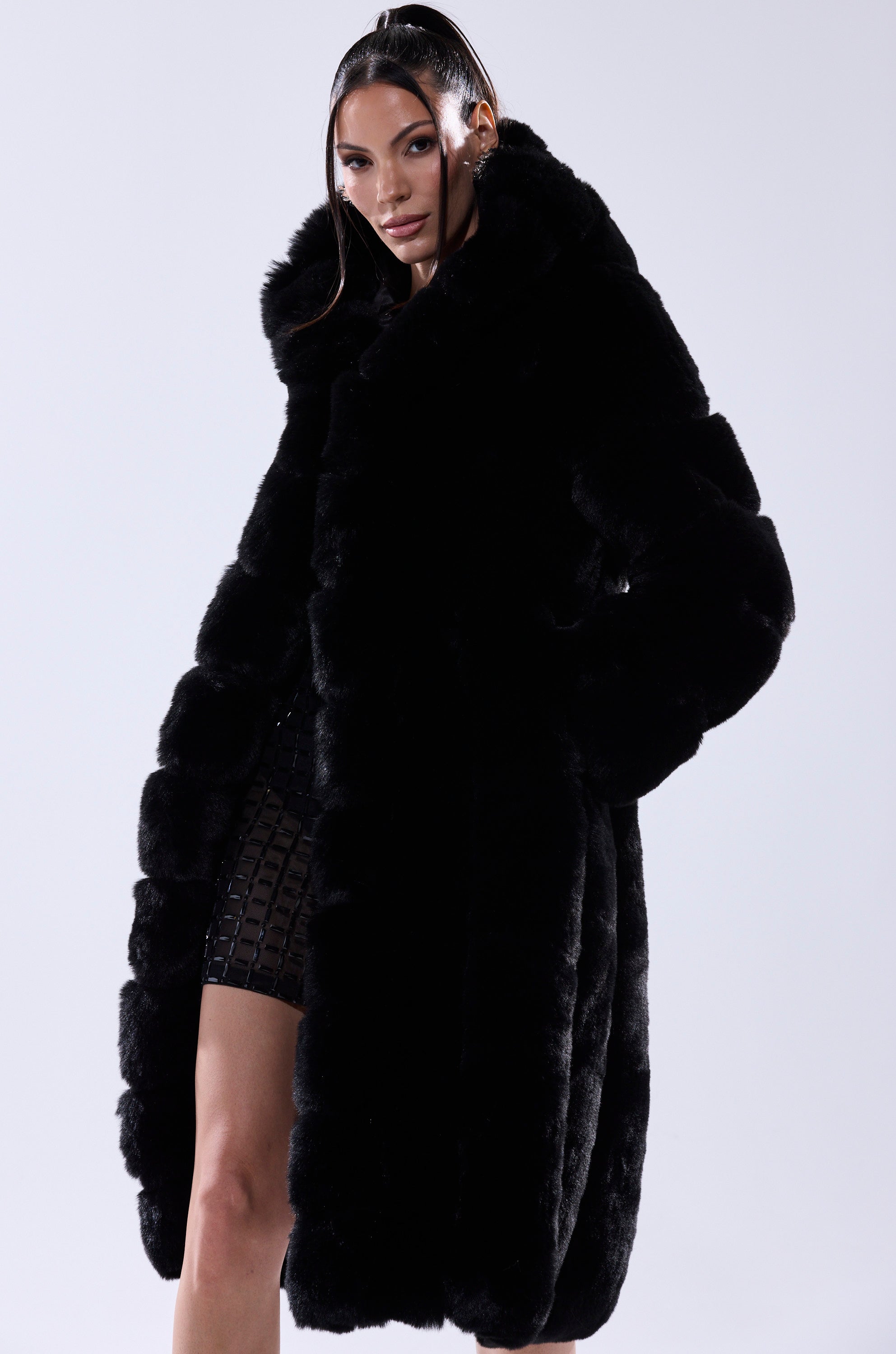 A woman with slicked-back hair wears the BERLIN HOODED FAUX FUR IN BLACK over a black grid-patterned dress, standing against a plain light background.