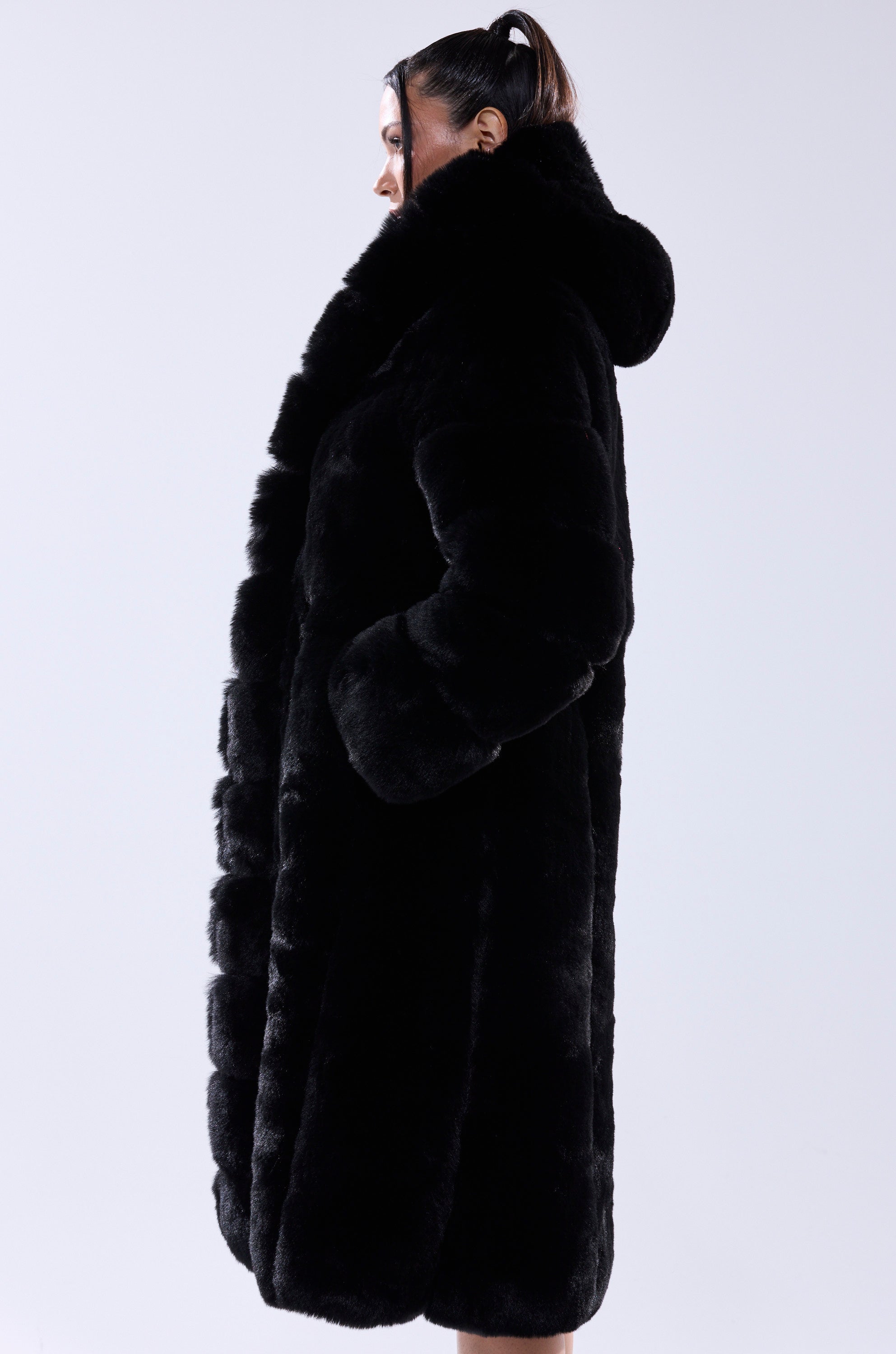 A person with dark hair in a ponytail stands sideways against a plain white background, wearing the BERLIN HOODED FAUX FUR IN BLACK—a black longline faux fur jacket featuring a large collar and hood.