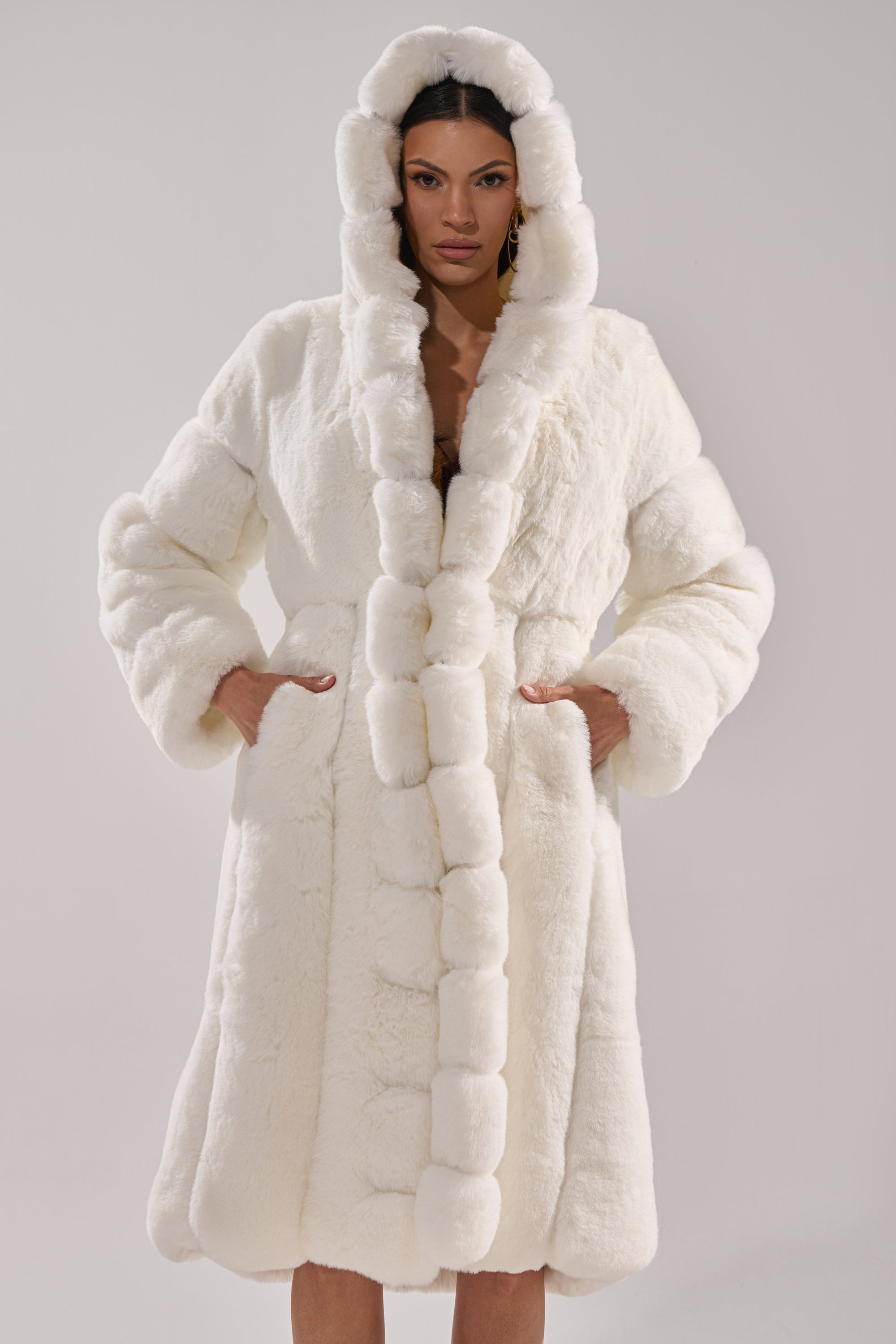 A woman stands with her hands in the pockets of the BERLIN HOODED FAUX FUR IN WHITE, a long, plush hooded faux fur coat, against a plain light background.