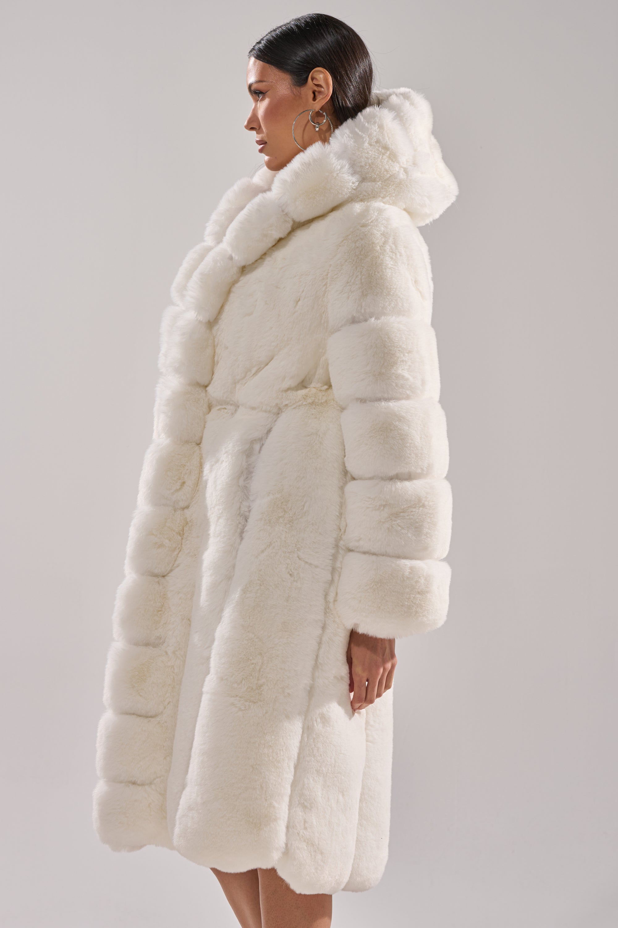 A woman in profile models the BERLIN HOODED FAUX FUR IN WHITE, featuring wide plush sleeves and a belted waist, set against a light gray background.