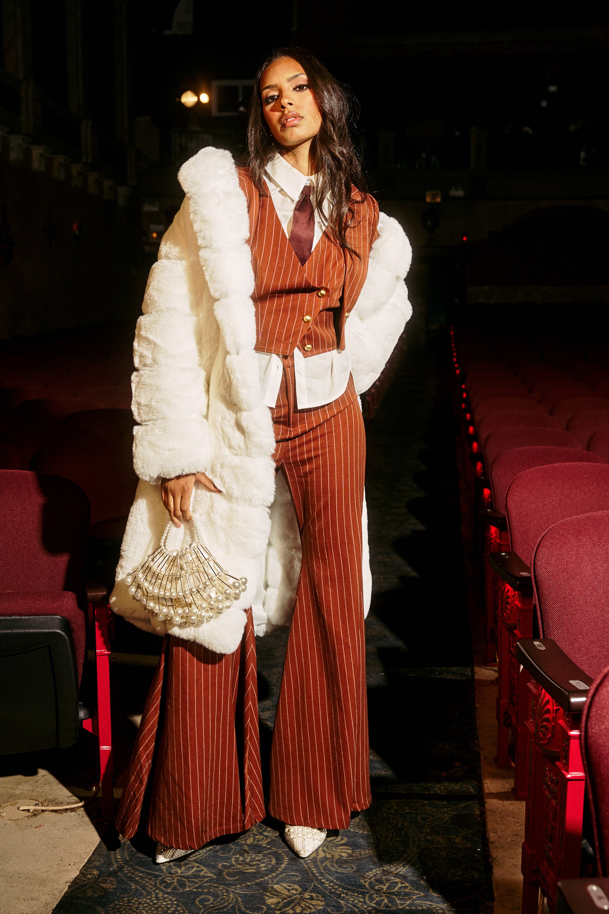 A woman stands in a theater aisle wearing a rust-colored pinstripe suit with flared pants, brown tie, pointed shoes, beaded handbag, and the BERLIN HOODED FAUX FUR IN WHITE draped over her shoulders.