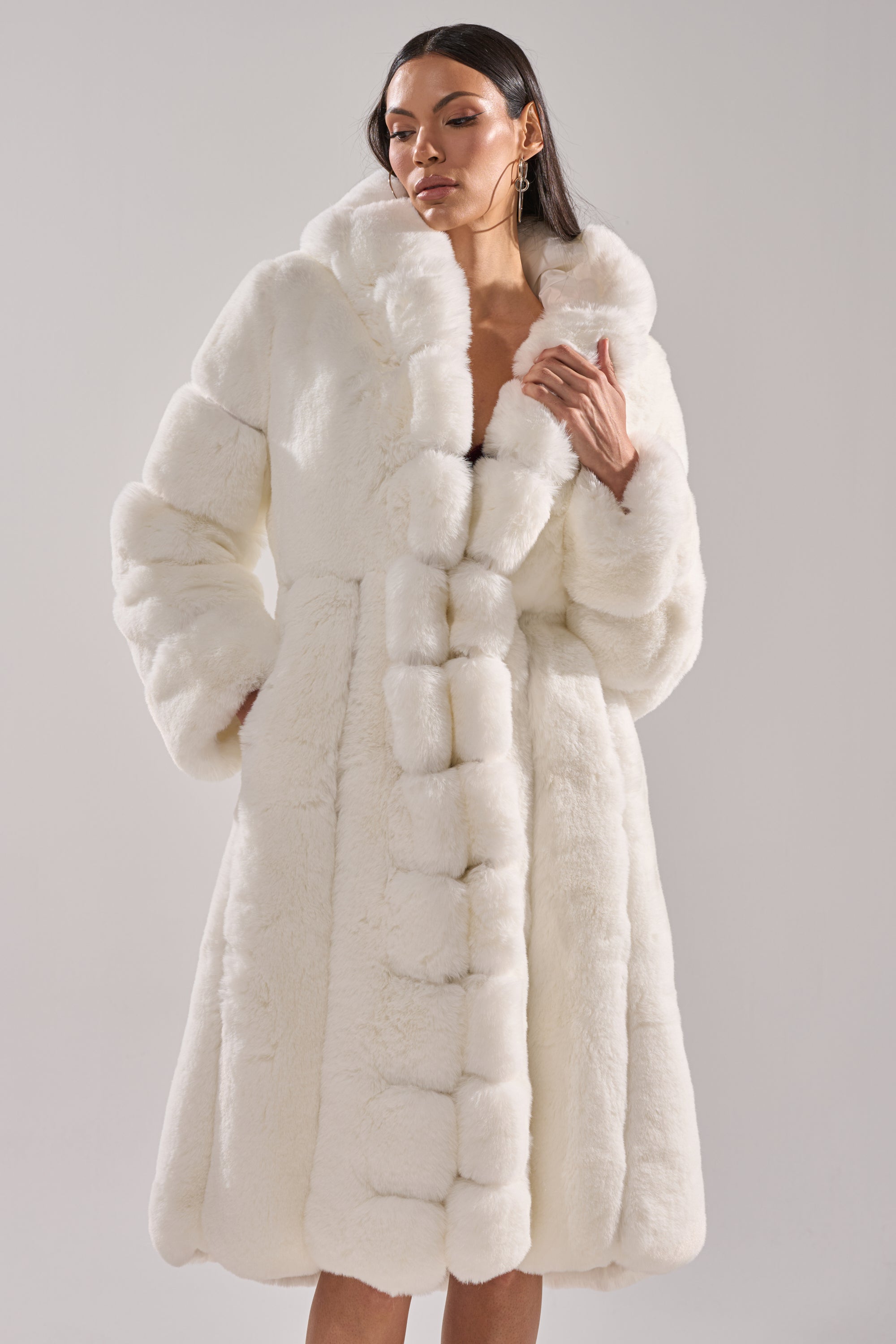 A woman with long dark hair wears the BERLIN HOODED FAUX FUR IN WHITE against a light grey background, exuding calm elegance—perfect stylish and cozy winter outerwear.