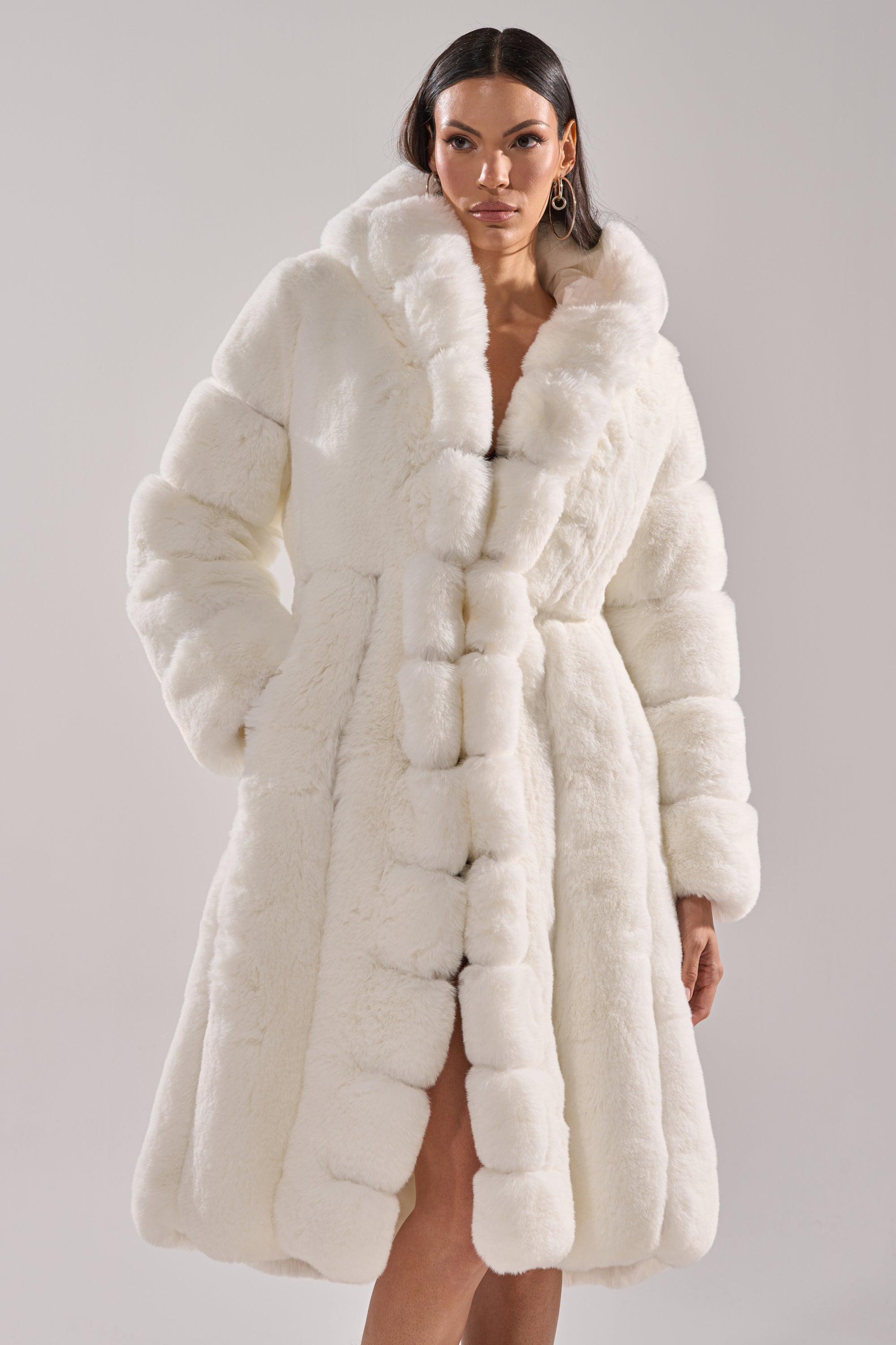 A woman models the BERLIN HOODED FAUX FUR IN WHITE—a plush, textured faux fur coat with a hood. This elegant winter outerwear falls just above her knees, offering both style and warmth against a plain light background.