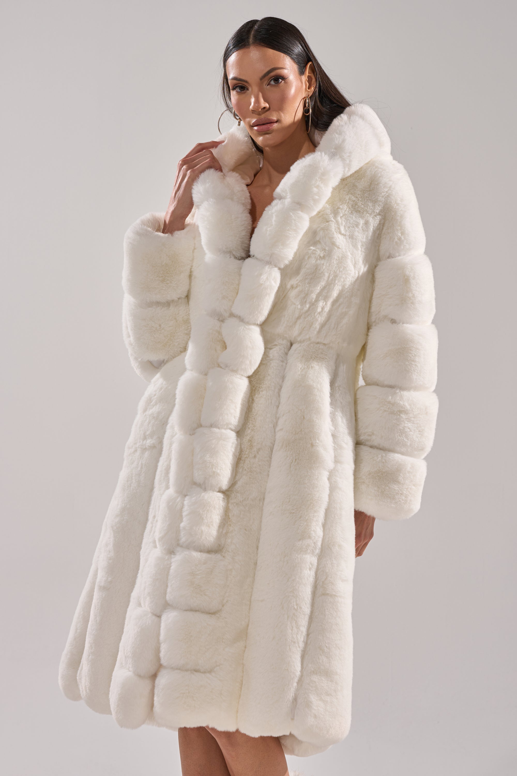 A woman models the BERLIN HOODED FAUX FUR IN WHITE, a luxurious long coat with a wide collar and panel detailing, posing against a plain light background to showcase refined winter style.