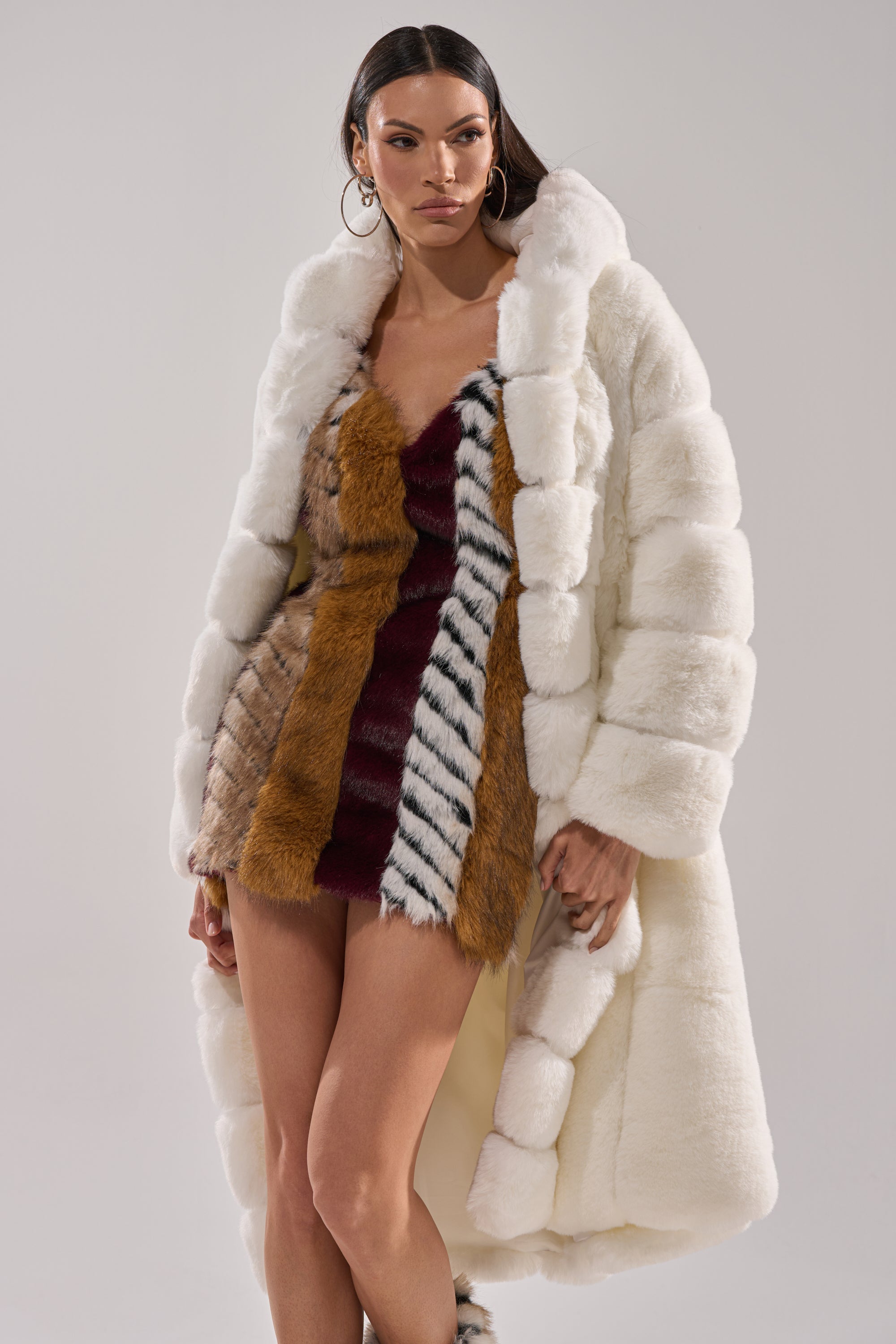 A woman models the BERLIN HOODED FAUX FUR IN WHITE—a luxurious long white faux fur coat—over a short, multicolored fur dress with brown, burgundy, and striped accents. She accessorizes with large hoop earrings and sleek, straight hair.