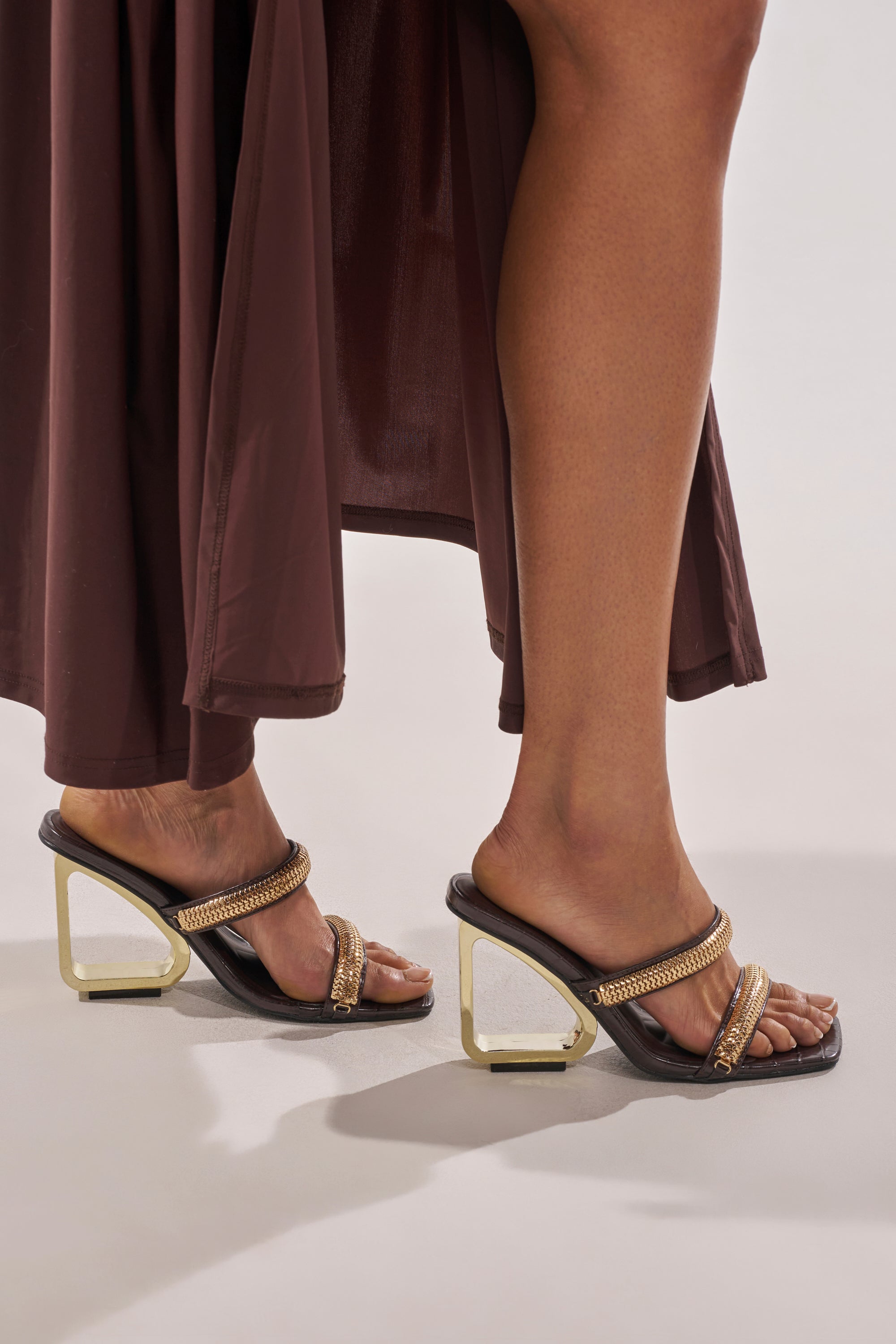 A person wearing a brown skirt and the AZALEA WANG BESSIE CHOCOLATE CROC GOLD EMBELLISHED WEDGE SANDAL, featuring gold hardware straps and unique geometric gold heels, stands against a plain light background. 1
