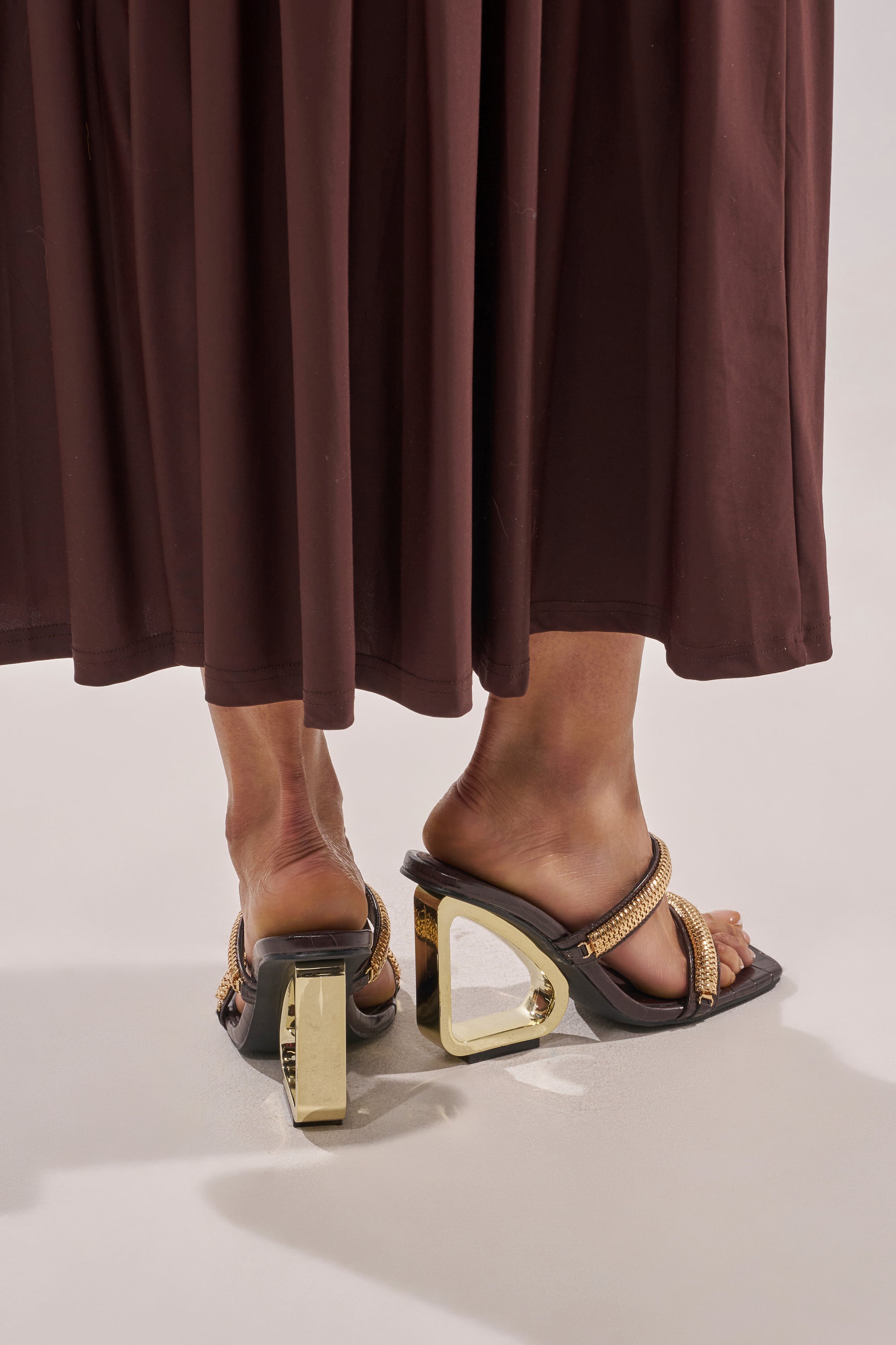 A person wears a long brown skirt and AZALEA WANG BESSIE CHOCOLATE CROC GOLD EMBELLISHED WEDGE SANDALS featuring gold hardware straps and unique geometric triangle-shaped gold heels. 3