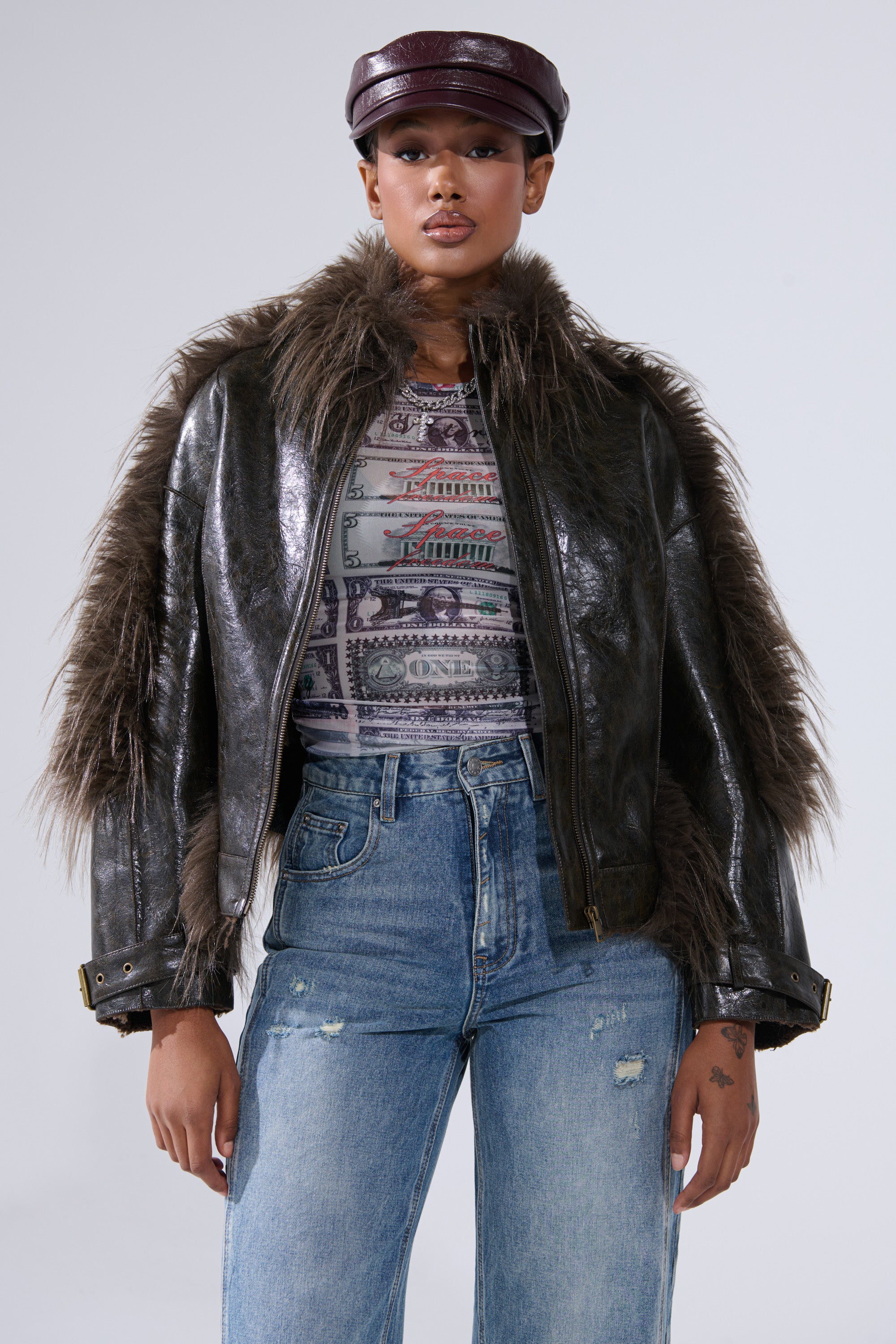 A woman in the VOODOO FUR TRIM MOTO jacket, paired with a patterned top, high-waisted ripped jeans, and a maroon cap stands against a light background, exuding biker-chic streetwear style.