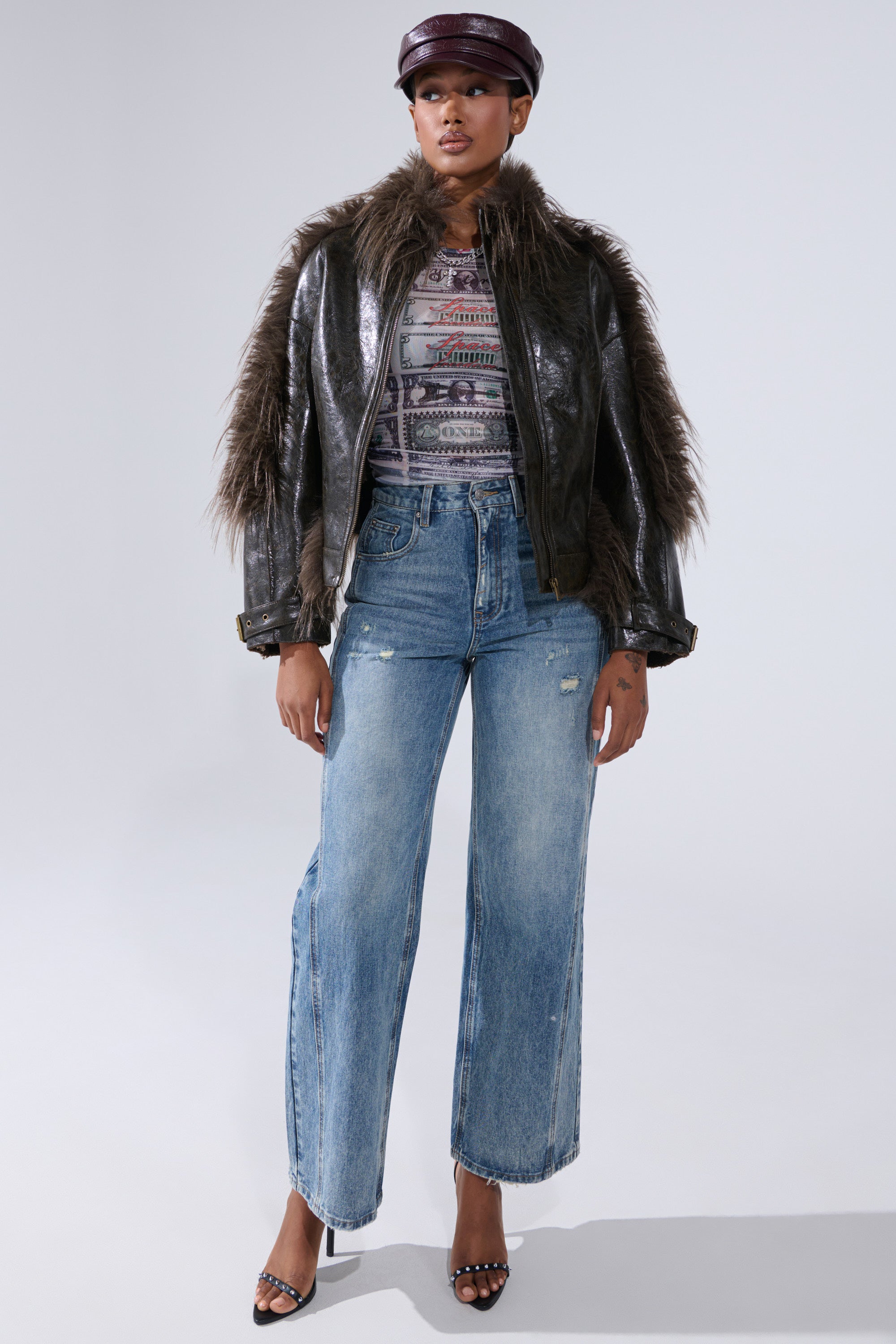 A person stands confidently in the VOODOO FUR TRIM MOTO biker-chic jacket, paired with a patterned top, wide-leg blue jeans, black strappy heels, and a maroon leather cap against a plain light background.