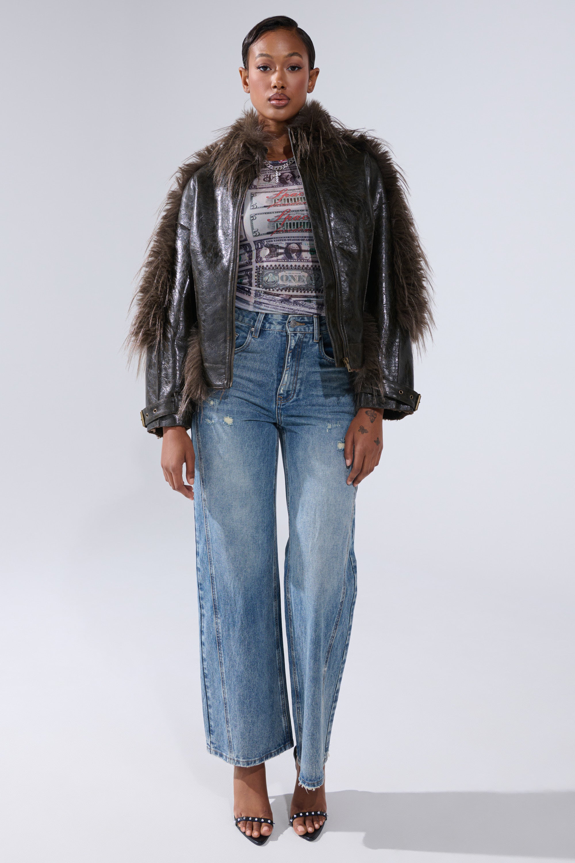 A woman faces forward against a plain light background, wearing the VOODOO FUR TRIM MOTO jacket, a patterned top, wide-leg blue jeans, and black heeled sandals.