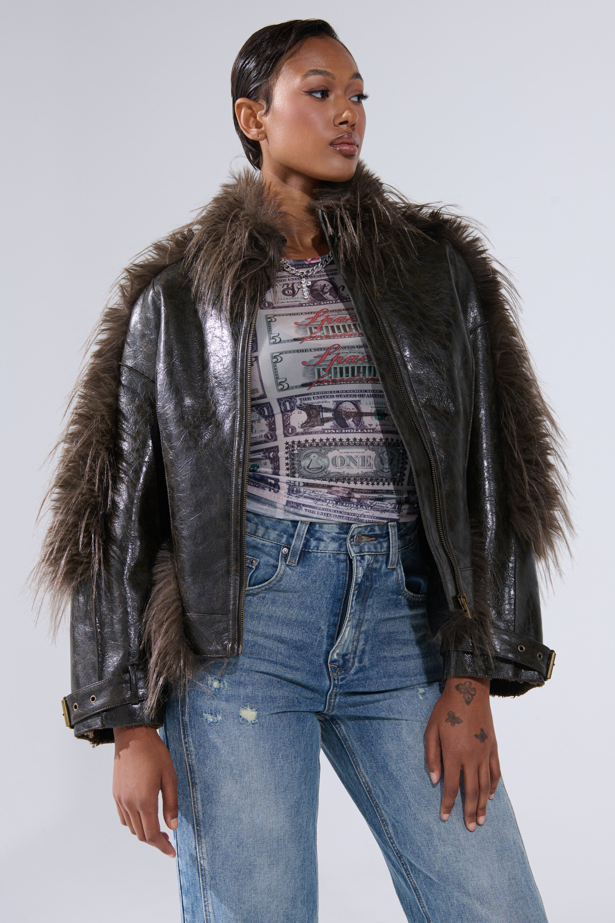 A woman wears the VOODOO FUR TRIM MOTO jacket—shiny dark faux leather with a shaggy fur collar—paired with a patterned top and high-waisted blue jeans, standing confidently against a plain light background.