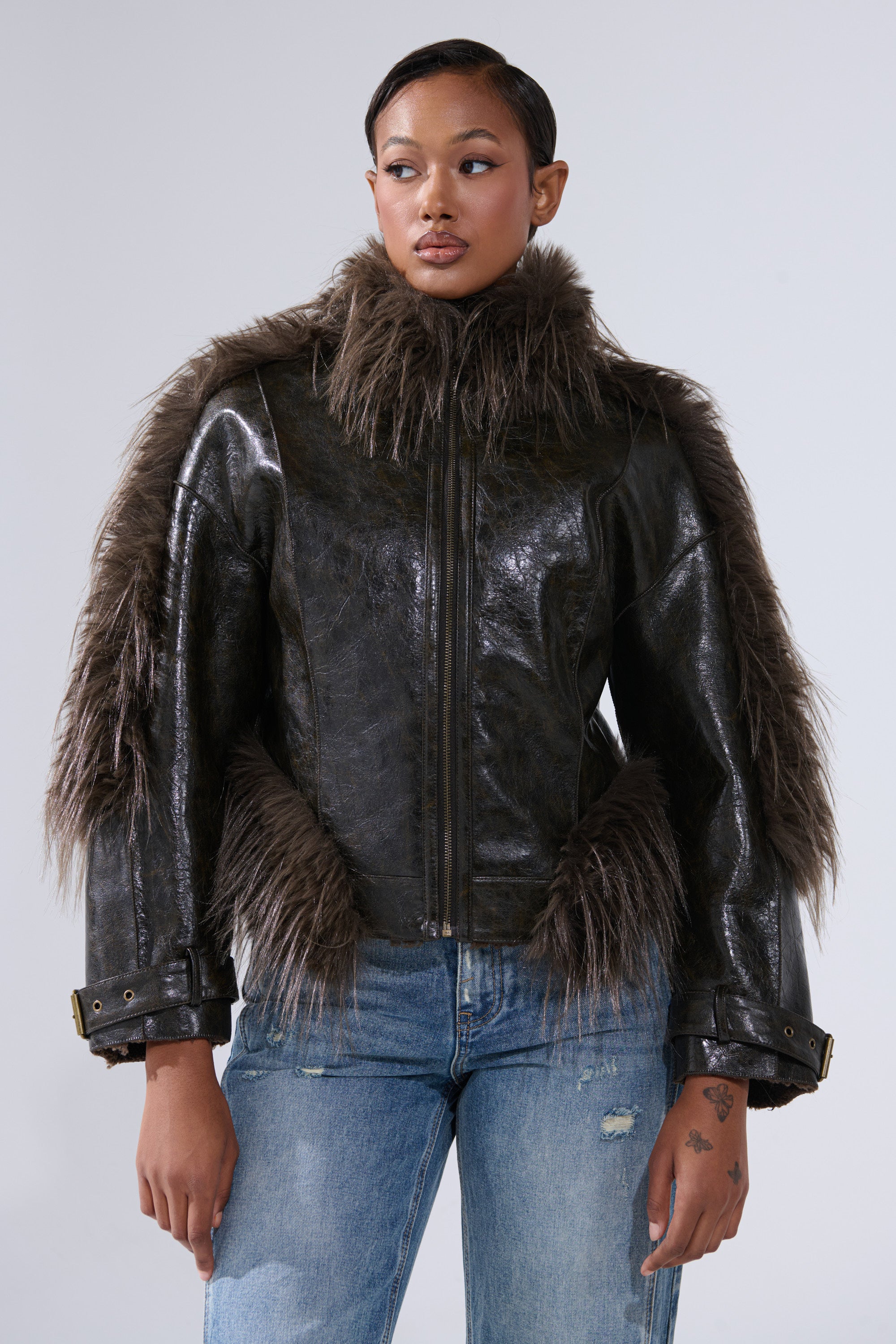 A woman models the VOODOO FUR TRIM MOTO, a shiny black faux leather jacket with brown fur collar, shoulder, and cuff trim, styled with light blue ripped jeans against a plain light gray background.