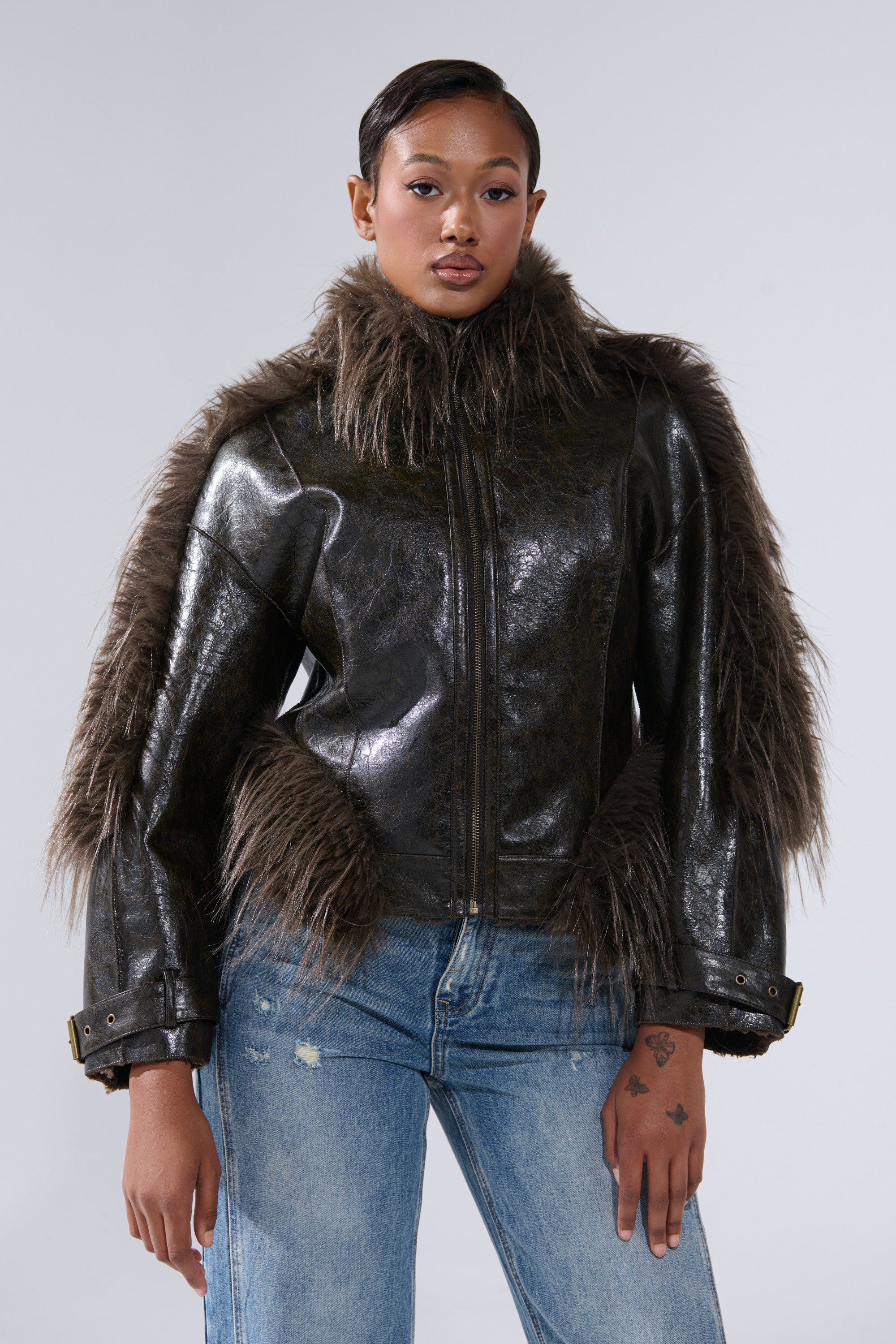 Wearing the VOODOO FUR TRIM MOTO, a shiny faux leather jacket with shaggy brown fur on collar, sleeves, and sides, paired with blue distressed jeans for a biker-chic streetwear look against a plain gray background.