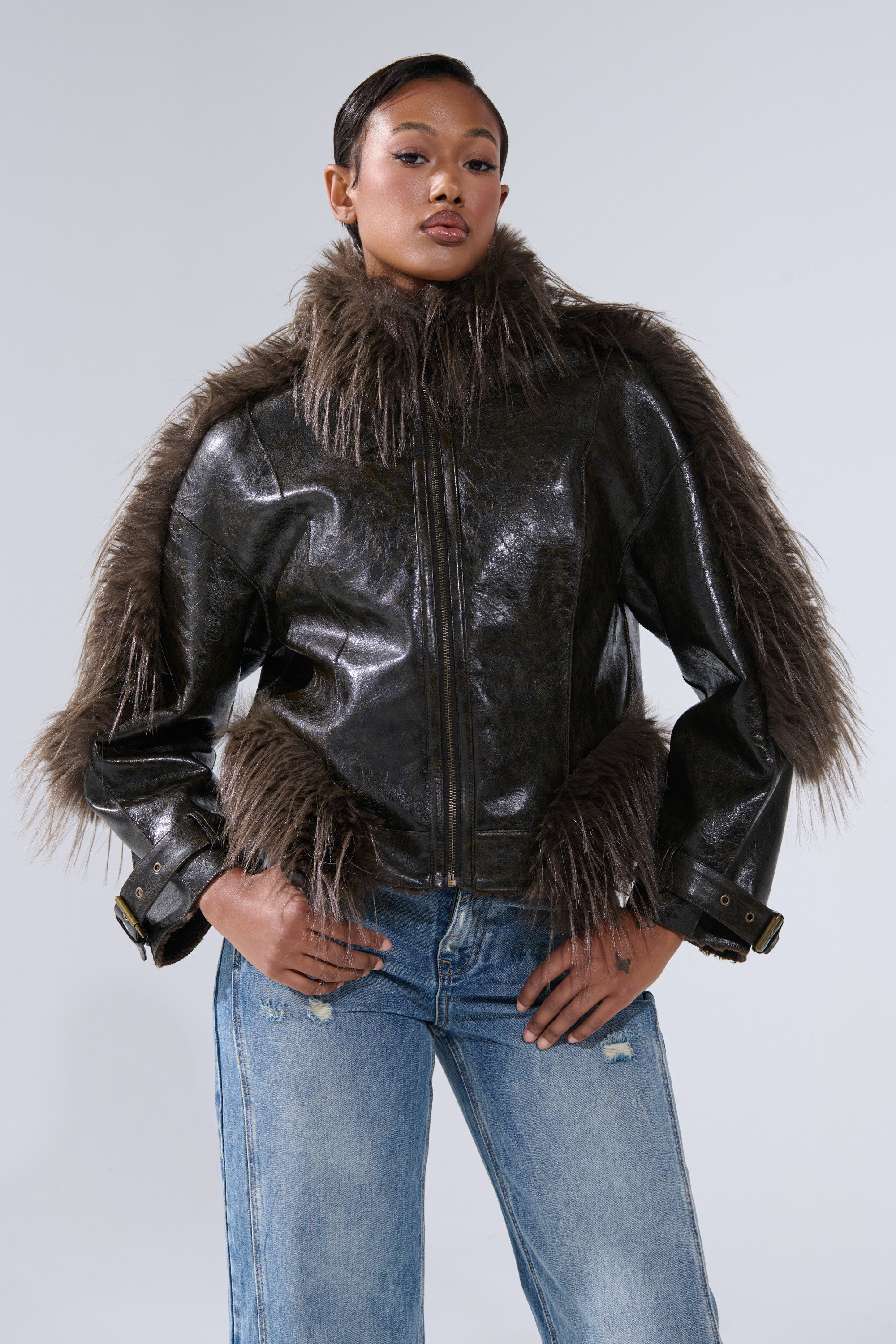 A woman stands confidently in the VOODOO FUR TRIM MOTO, a shiny black faux leather moto jacket with large brown fur trim, paired with blue ripped jeans against a plain light gray background.
