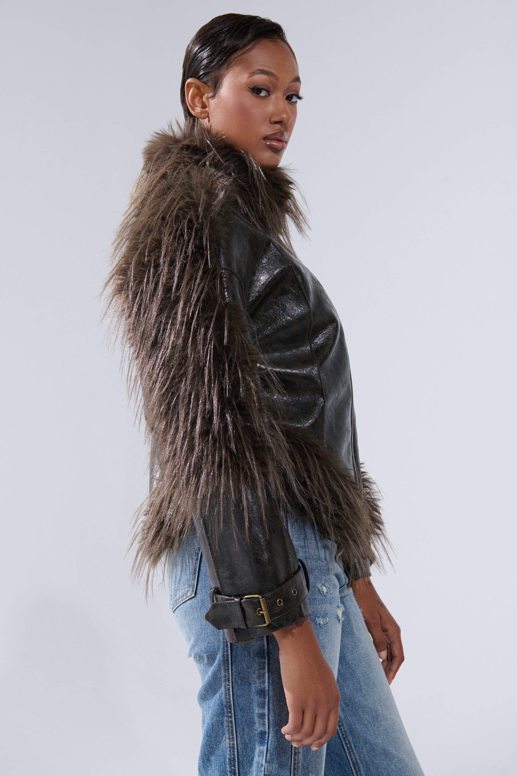 A woman stands sideways wearing the VOODOO FUR TRIM MOTO jacket with thick, shaggy fur trim and blue jeans, looking confidently over her shoulder against a plain background.