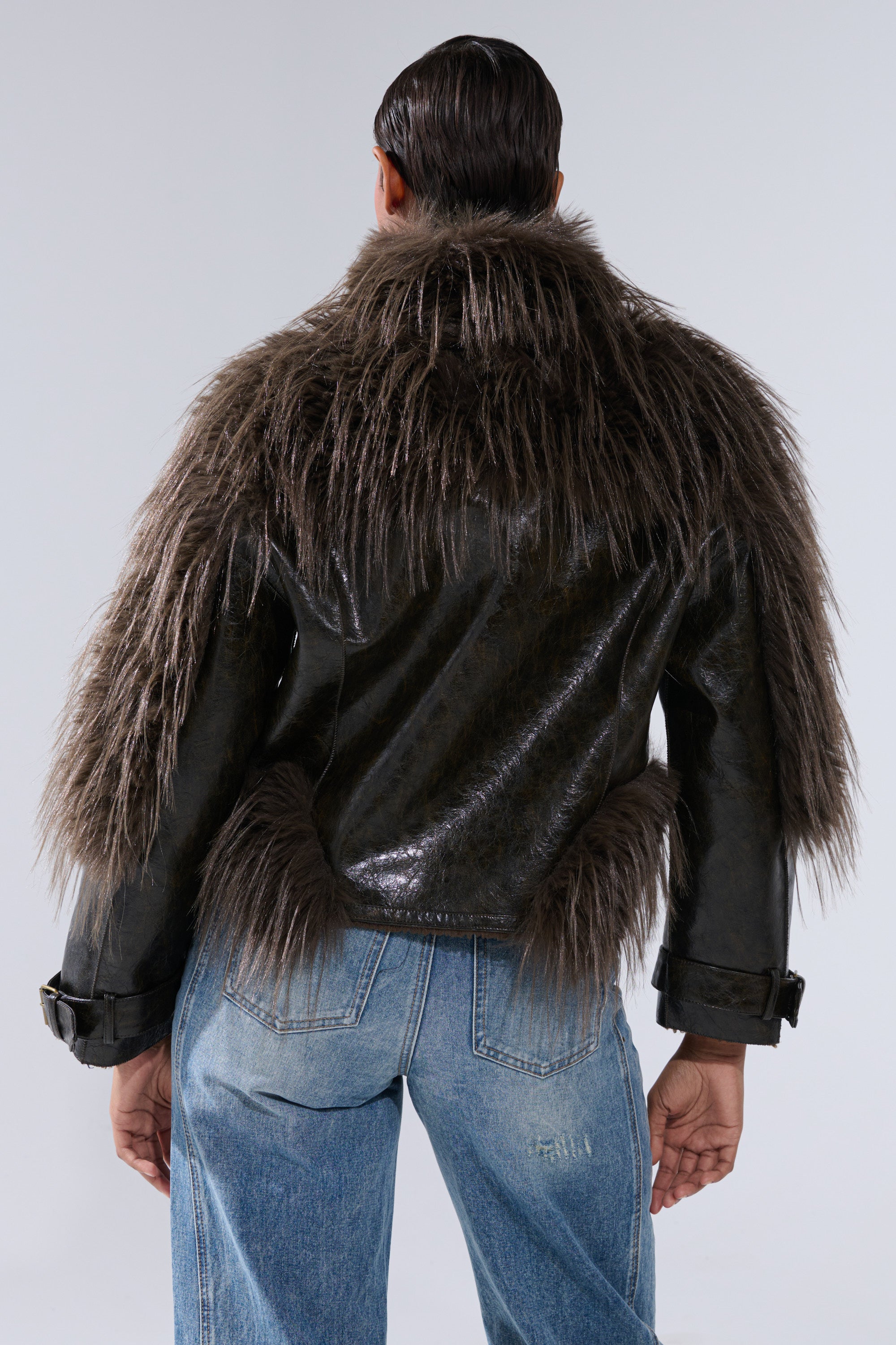 A person faces away, wearing the VOODOO FUR TRIM MOTO—a shiny black jacket with thick, long brown faux fur on the shoulders, upper back, and lower waist—paired with light blue jeans against a plain light background.