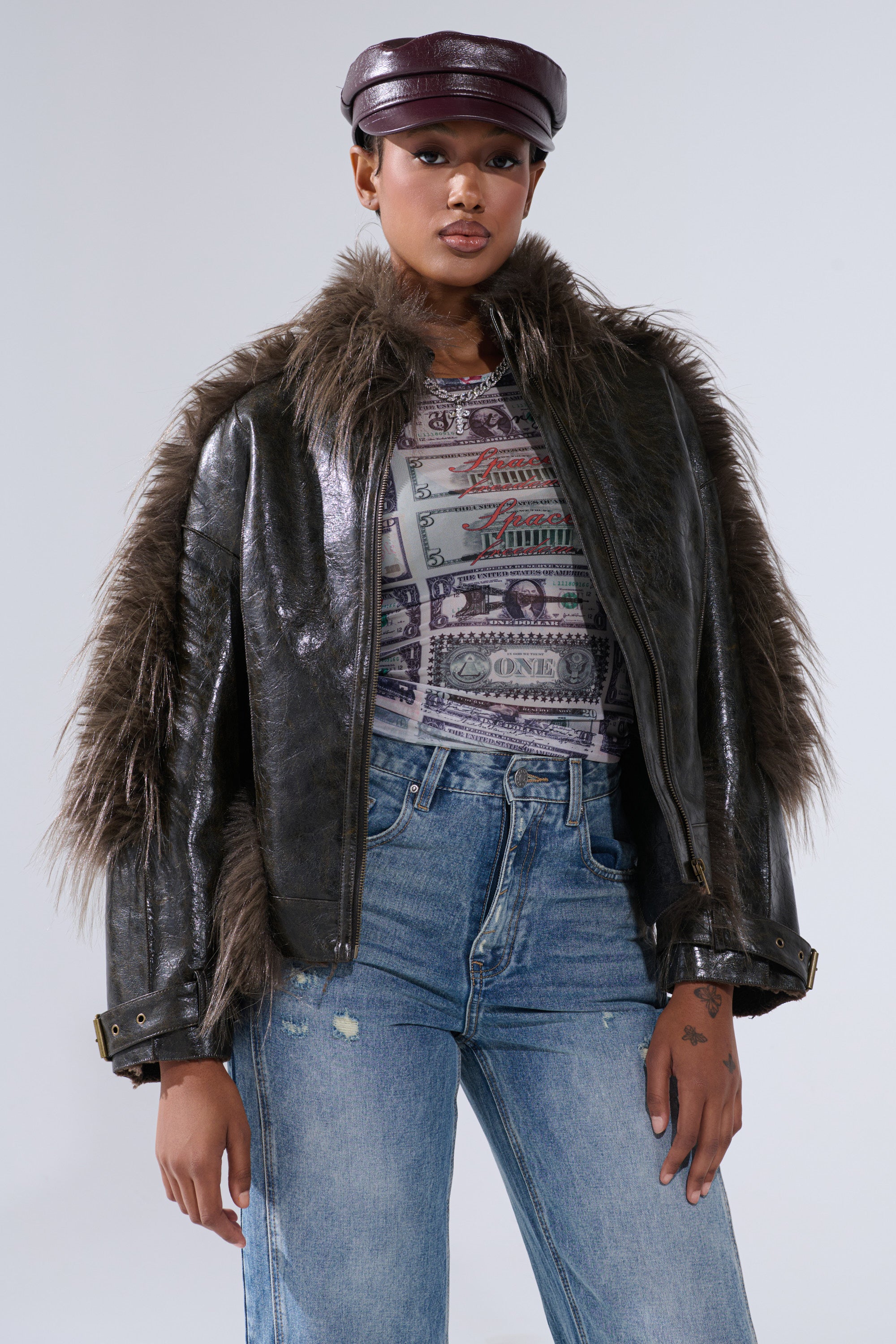 A person poses confidently against a plain light background, styled in the VOODOO FUR TRIM MOTO jacket paired with a printed shirt, ripped high-waisted jeans, and a maroon leather cap.