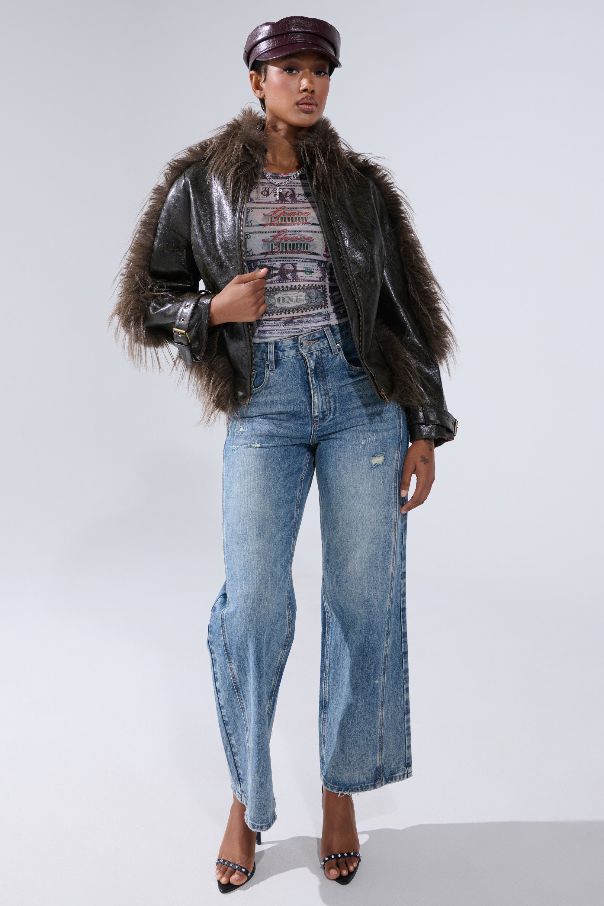 Wearing the VOODOO FUR TRIM MOTO jacket, a patterned top, wide-leg light blue jeans, black heeled sandals, and a dark leather newsboy cap, a person stands against a plain light gray background with biker-chic streetwear flair.