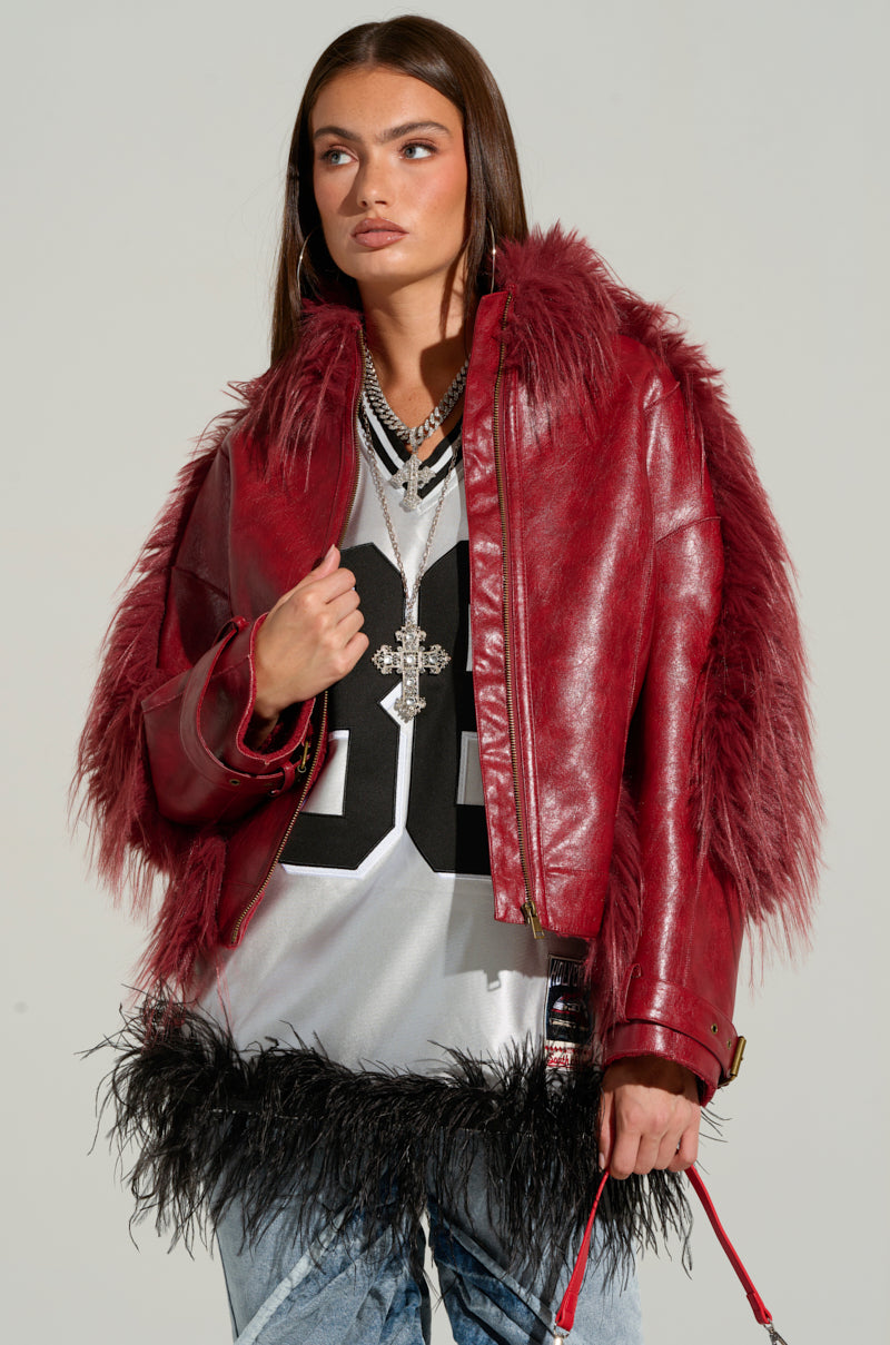 A person with long hair stands confidently, wearing the striking OVERZEALOUS FUR TRIM BOMBER with a red, fur-trimmed lining over a black and white sports jersey emblazoned with the number "23." Their look is completed with layered necklaces and feather-accented grey pants.