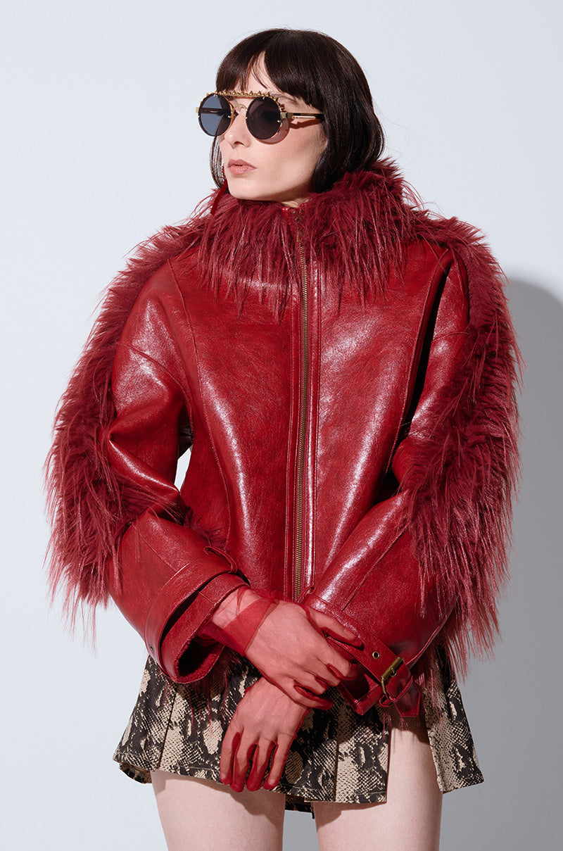 A woman stands against a plain background, wearing round sunglasses and the OVERZEALOUS FUR TRIM BOMBER, a distinctive red leather jacket with faux sherpa lining on the shoulders and sleeves. She pairs it with a snakeskin-patterned mini skirt and poses with her gloved hands touching.
