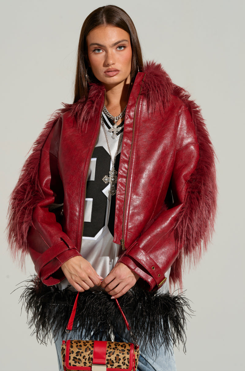 A woman with long straight hair wears the Overzealous Fur Trim Bomber, which features a vibrant red fur trim, over a grey shirt adorned with a large black number. She holds a small leopard print purse highlighted by red accents and embellished with black feathers. She stands against a plain background, gazing directly at the camera.