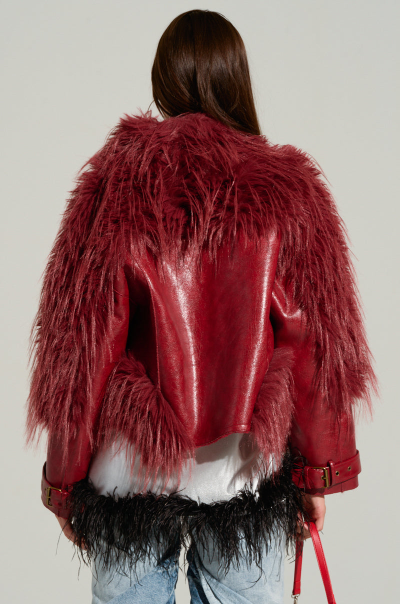 A person with long brown hair is wearing the OVERZEALOUS FUR TRIM BOMBER, which features wide, shaggy fur trim on the back and sleeves and a textured, furry appearance. They are also dressed in light blue jeans and holding a small red object in their left hand.
