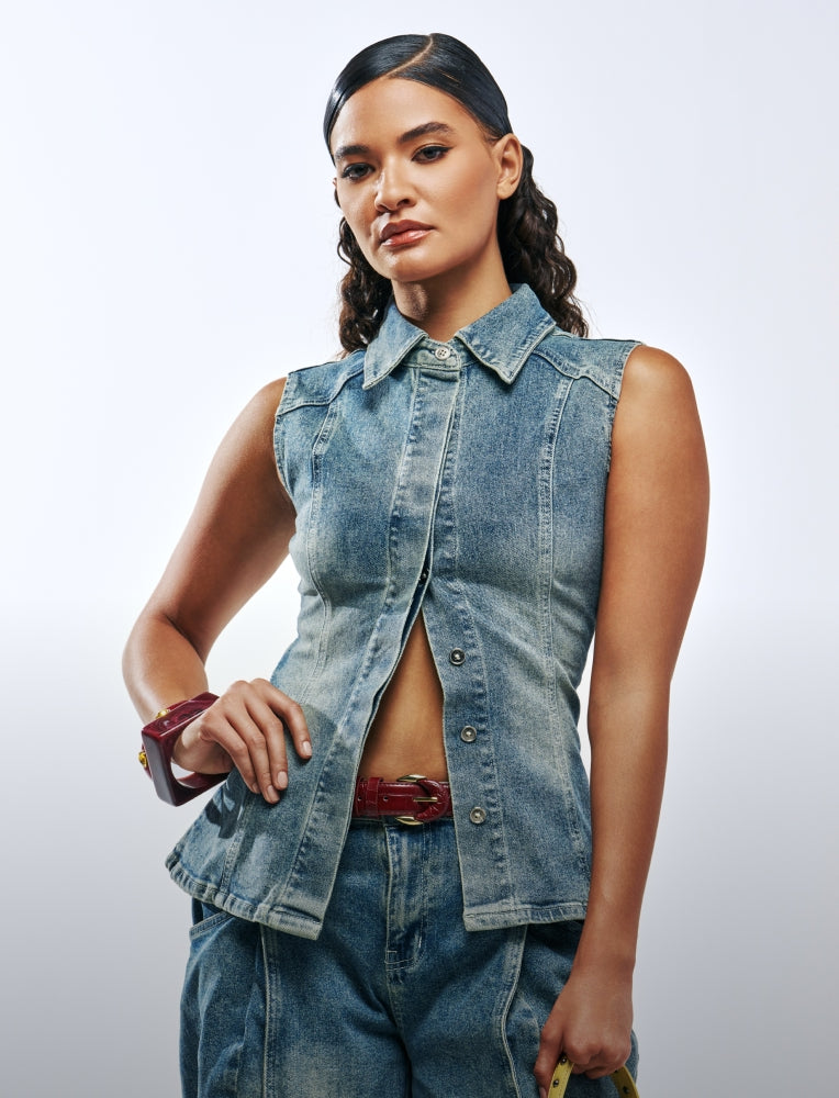 A woman modeling a sleeveless light-wash denim vest with a button-front closure and pointed collar, paired with matching denim bottoms, against a light gray studio background.