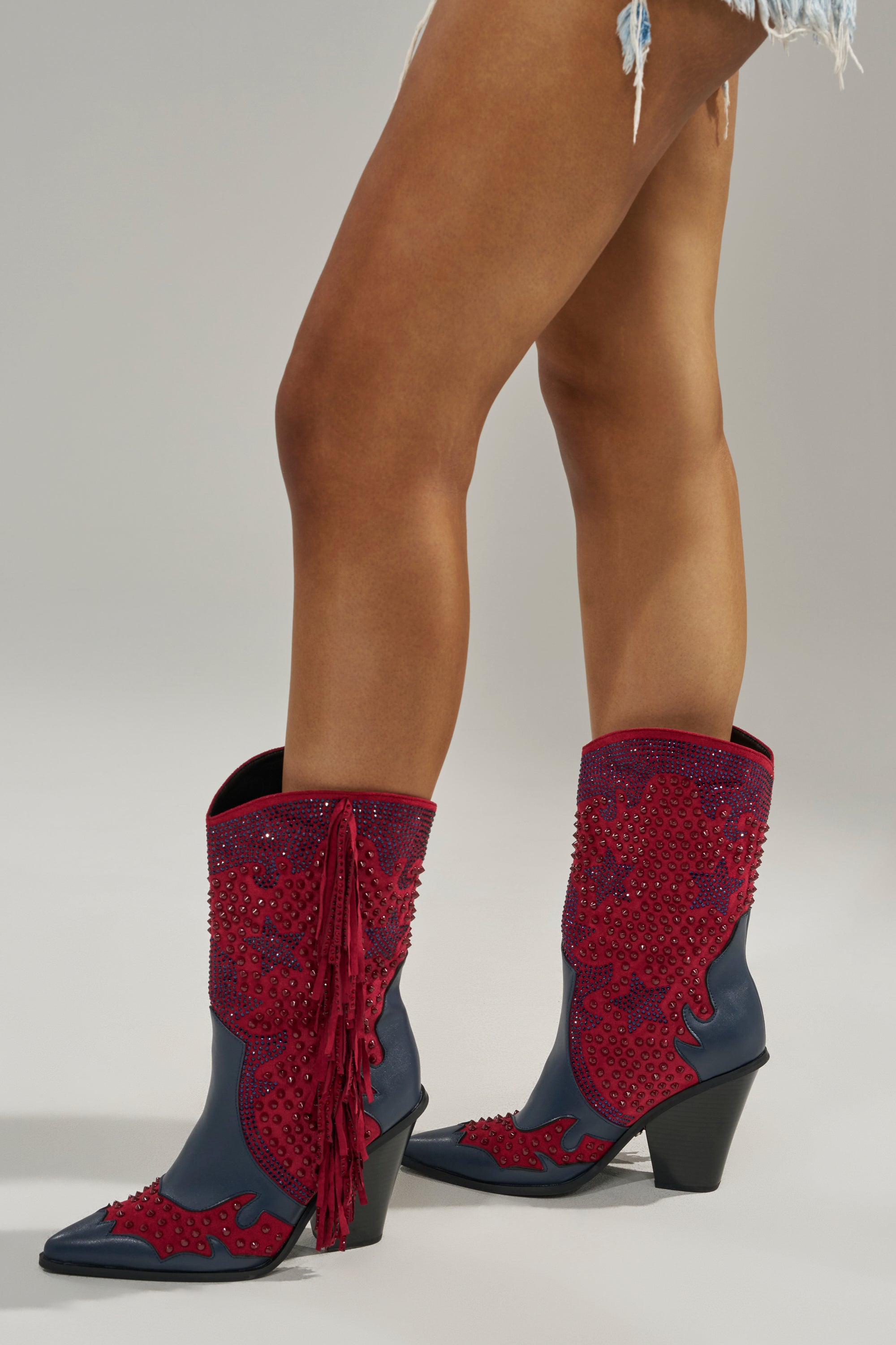 A person wearing frayed denim shorts and AZALEA WANG BETHANIE MULTI RHINESTONE FRINGE WESTERN BOOTIE stands on a neutral gray background.