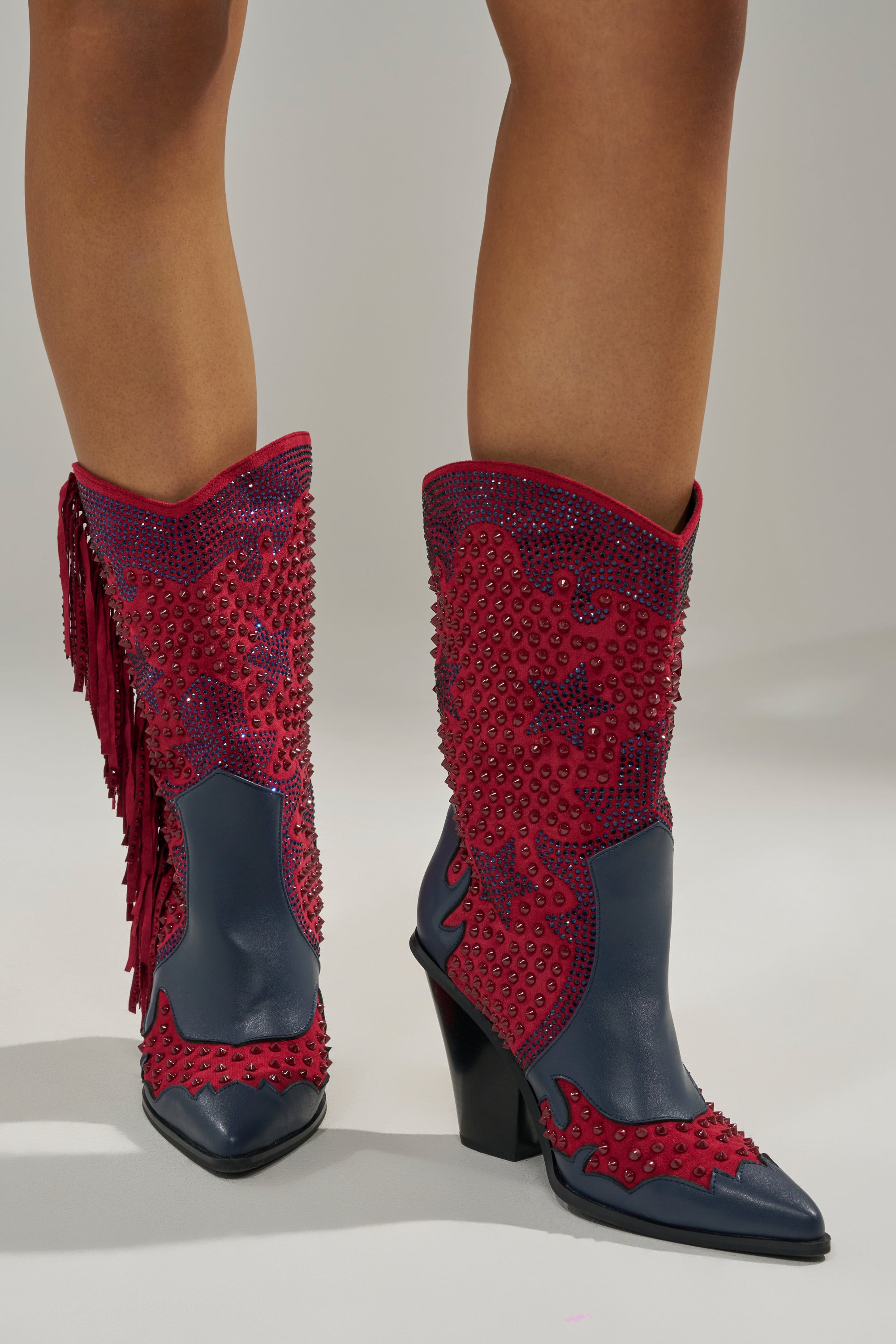Someone stands on a light floor wearing AZALEA WANG BETHANIE MULTI RHINESTONE FRINGE WESTERN BOOTIES in navy blue and red, adorned with red studs, fringe, pointed toes, and angled heels.
