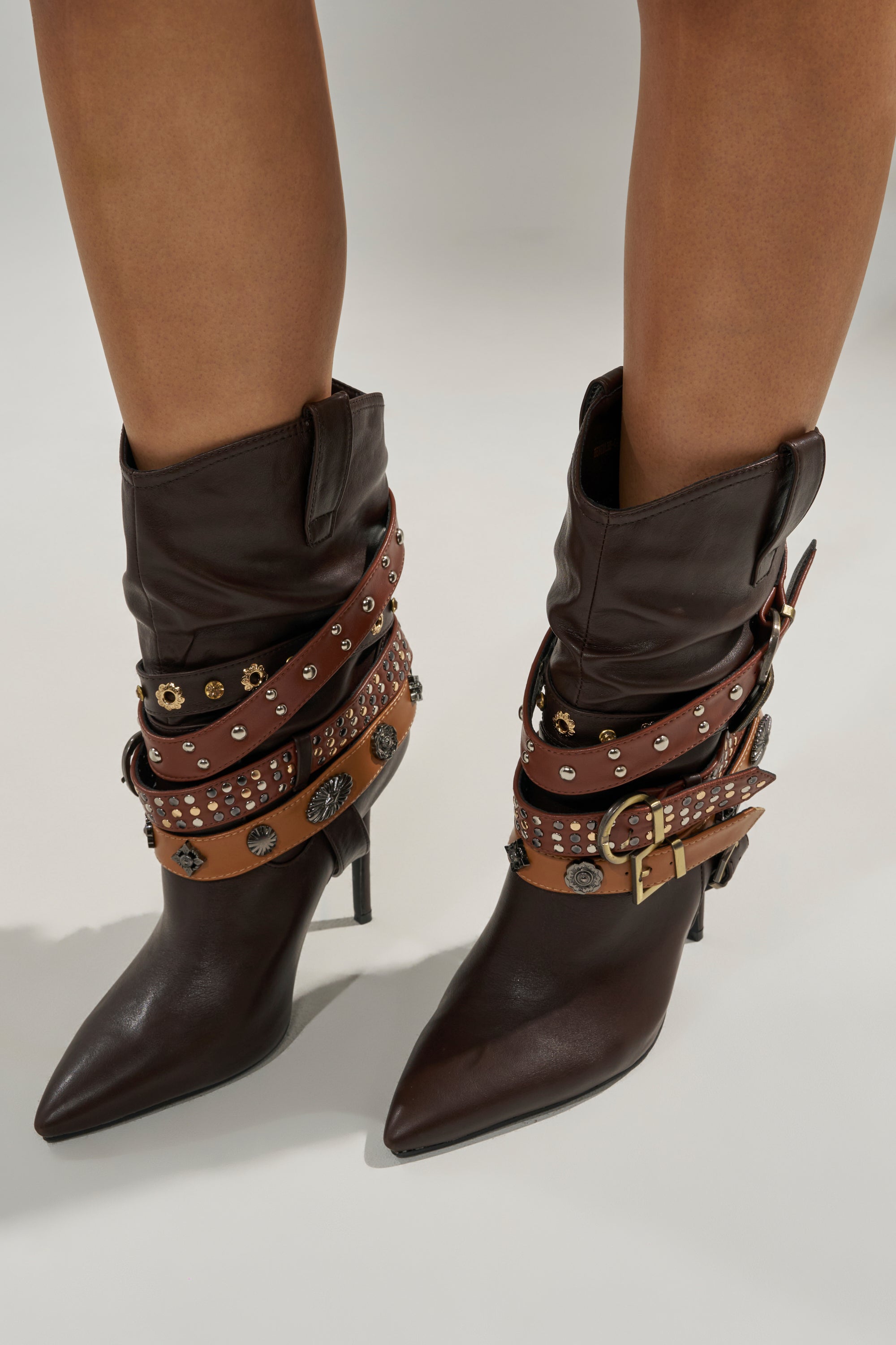 Close-up of a person wearing the AZALEA WANG BROWN STILETTO BOOTIE: dark brown pointed-toe heels featuring multiple leather straps, metal studs, conchos, and a bold gold buckle.