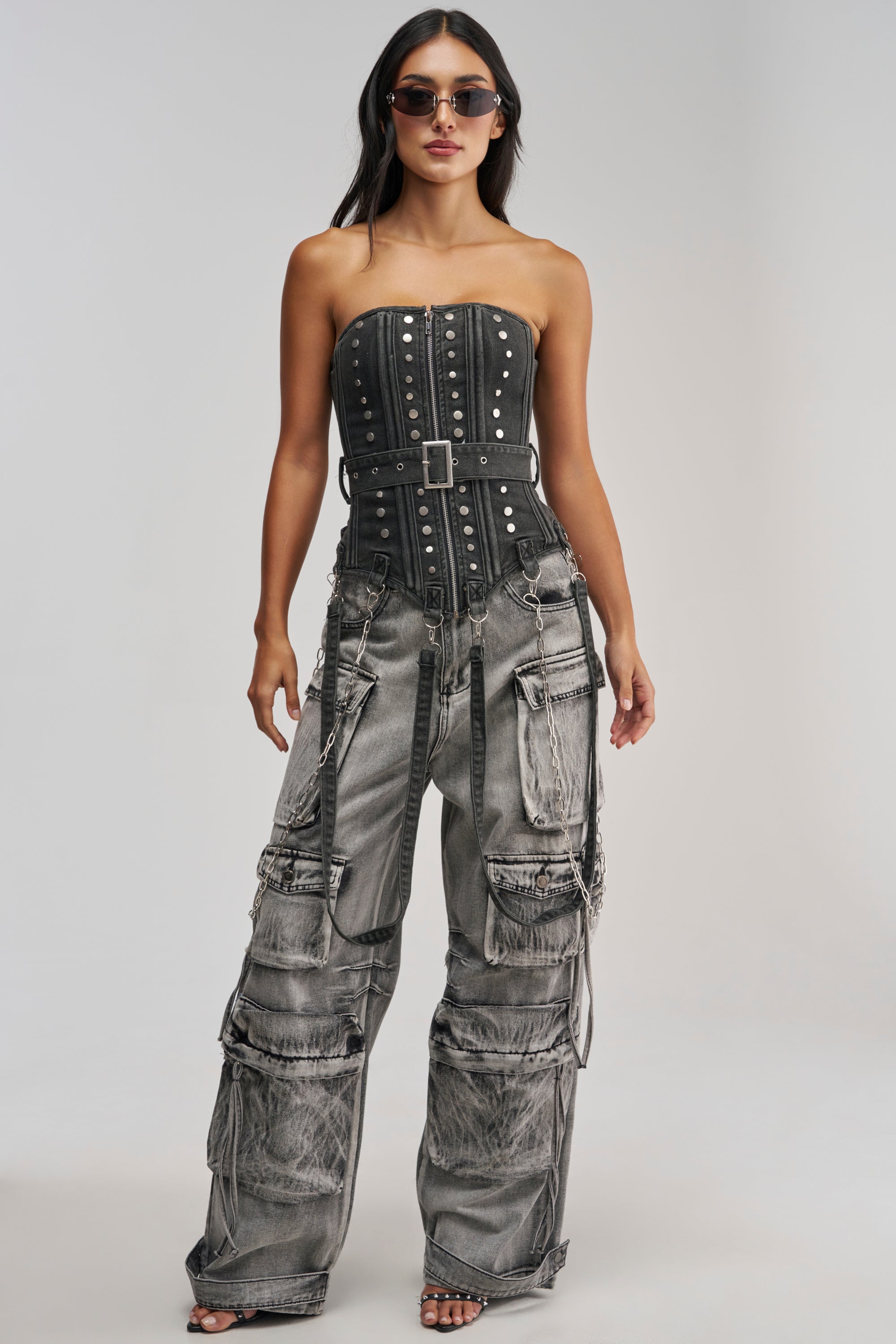 A woman wearing sunglasses and the PERK UP STUDDED CORSET IN DENIM, styled with a belt, paired with distressed wide-leg cargo jeans featuring multiple pockets and chains, stands against a plain light background.