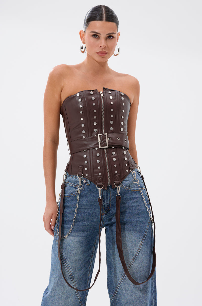 A woman with slicked-back hair models the PERK UP EMBELLISHED CORSET in brown leather, adorned with silver studs, belt, and chains. She pairs it with loose blue jeans and stands confidently against a plain white background.