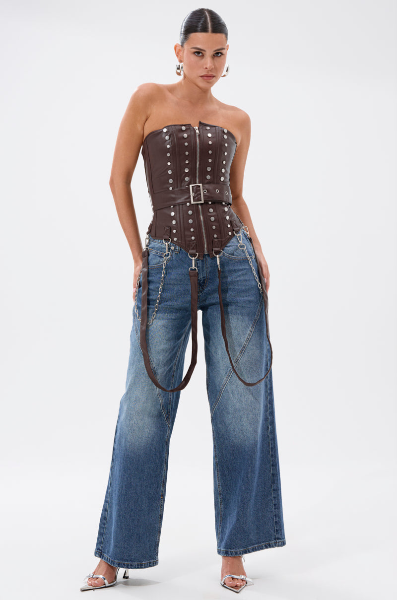 A woman stands against a white background wearing the PERK UP EMBELLISHED CORSET—a brown strapless top with silver studs—paired with wide-leg blue jeans with chain and strap details, silver heels, and hoop earrings.