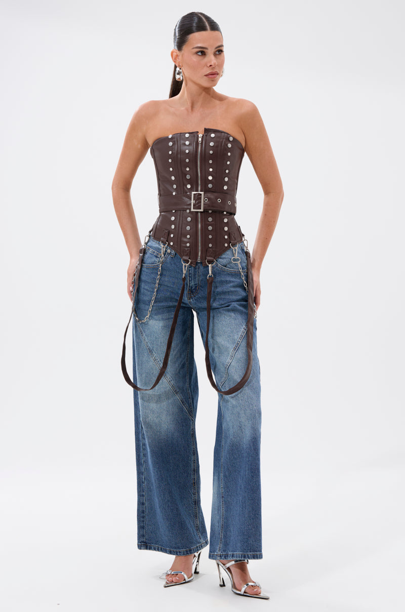 A woman stands against a white background wearing the PERK UP EMBELLISHED CORSET, a brown strapless faux leather top with metal studs, wide-leg blue jeans with chains and leather straps, and silver high-heeled sandals.
