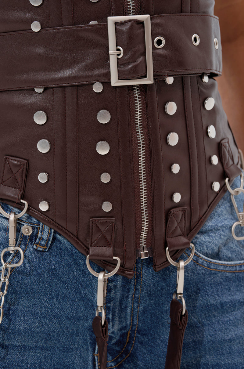 Close-up of someone wearing the PERK UP EMBELLISHED CORSET—brown faux leather with silver buckle, zipper, metal rings, and clip-on straps—styled with blue denim jeans.