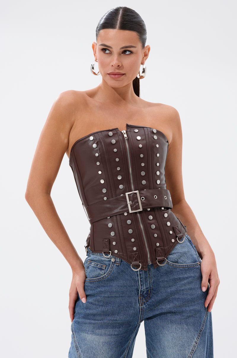 A woman with slicked-back hair wears the PERK UP EMBELLISHED CORSET—strapless faux leather with silver studs, zipper, and belt—paired with blue jeans. She looks to the side, hands in pockets, against a plain background.