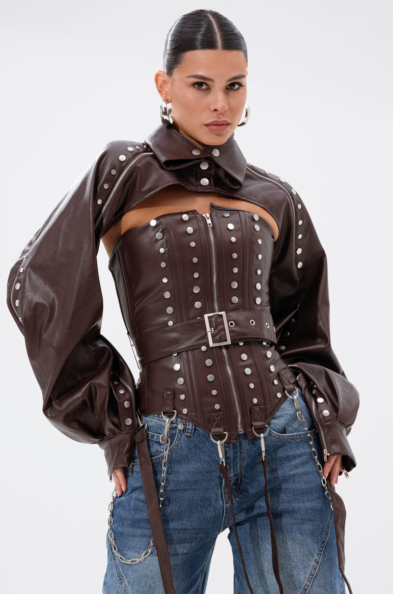 A woman with slicked-back hair wears the PERK UP EMBELLISHED CORSET—an embellished brown leather jacket with oversized sleeves—paired with blue jeans featuring silver chains, set against a plain white background.