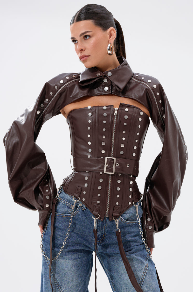 A woman wears the PERK UP EMBELLISHED CORSET in brown faux leather with silver studs, a matching cropped jacket with puffy sleeves, blue jeans, belts, zippers, chain accents, hoop earrings, and slicked-back hair.