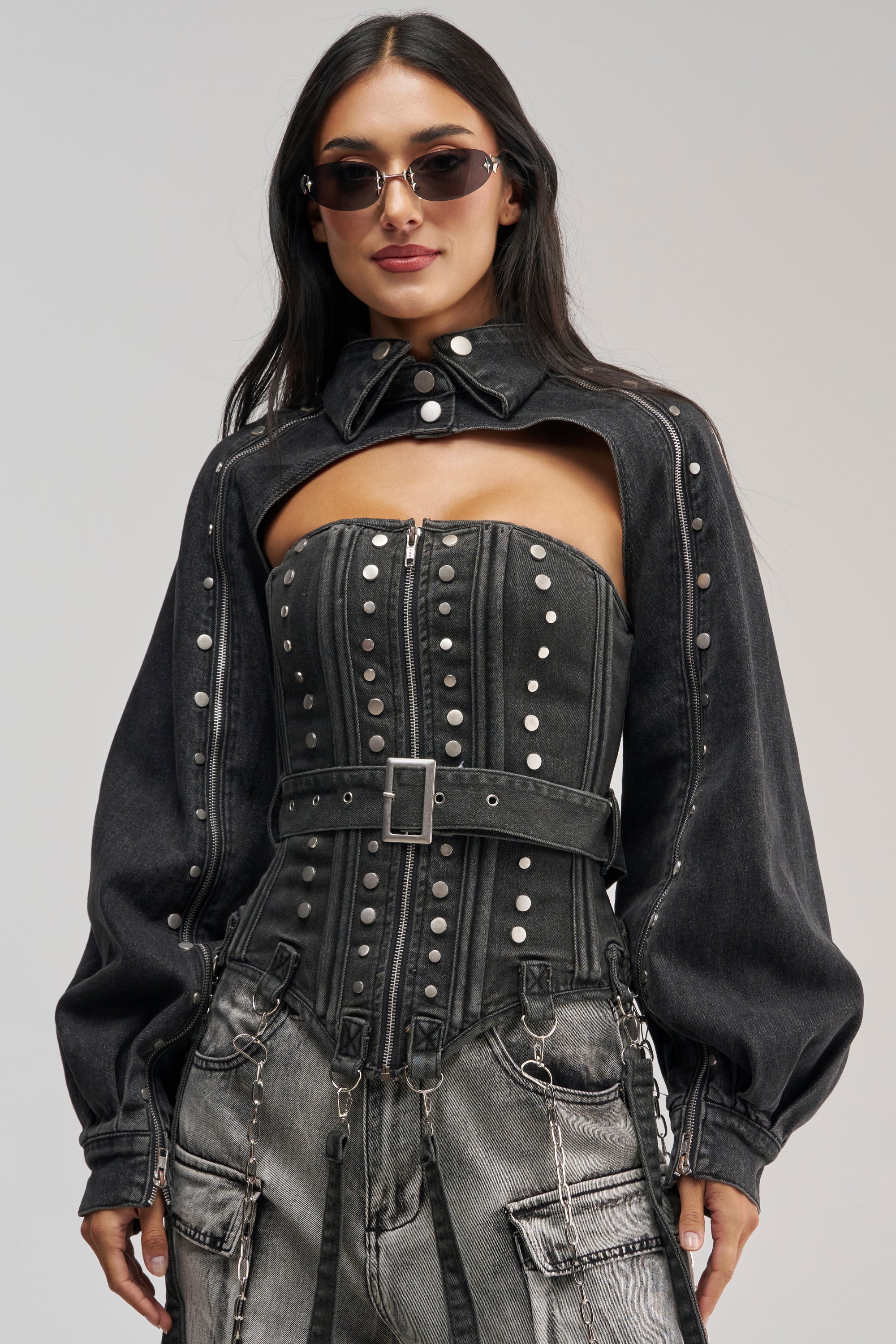 A woman poses confidently against a neutral background, wearing the PERK UP STUDDED BOLERO IN DENIM with puffed sleeves, paired with sunglasses, a studded bustier top featuring a zipper and belt, and high-waisted denim pants adorned with chains.