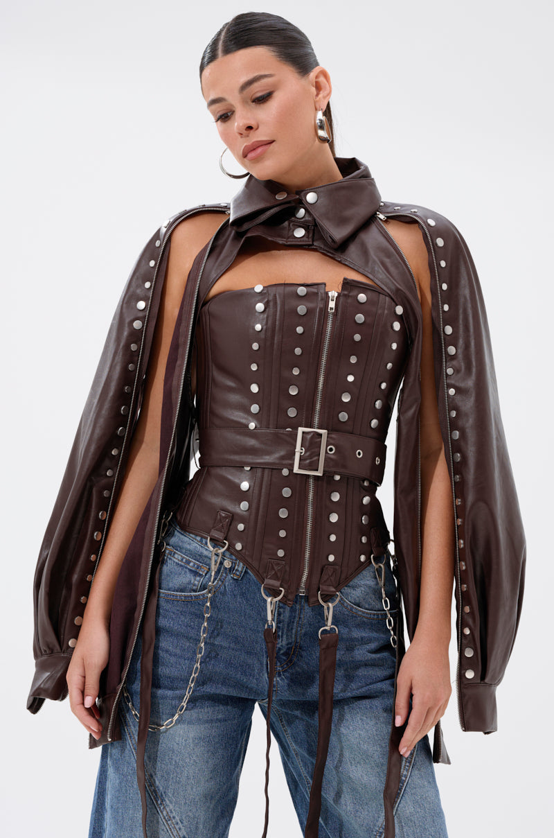 A woman wears the PERK UP EMBELLISHED BOLERO— a studded brown faux leather corset bolero with matching cape, belt, and chains— paired with blue jeans, hoop earrings, and slicked-back hair against a white background.