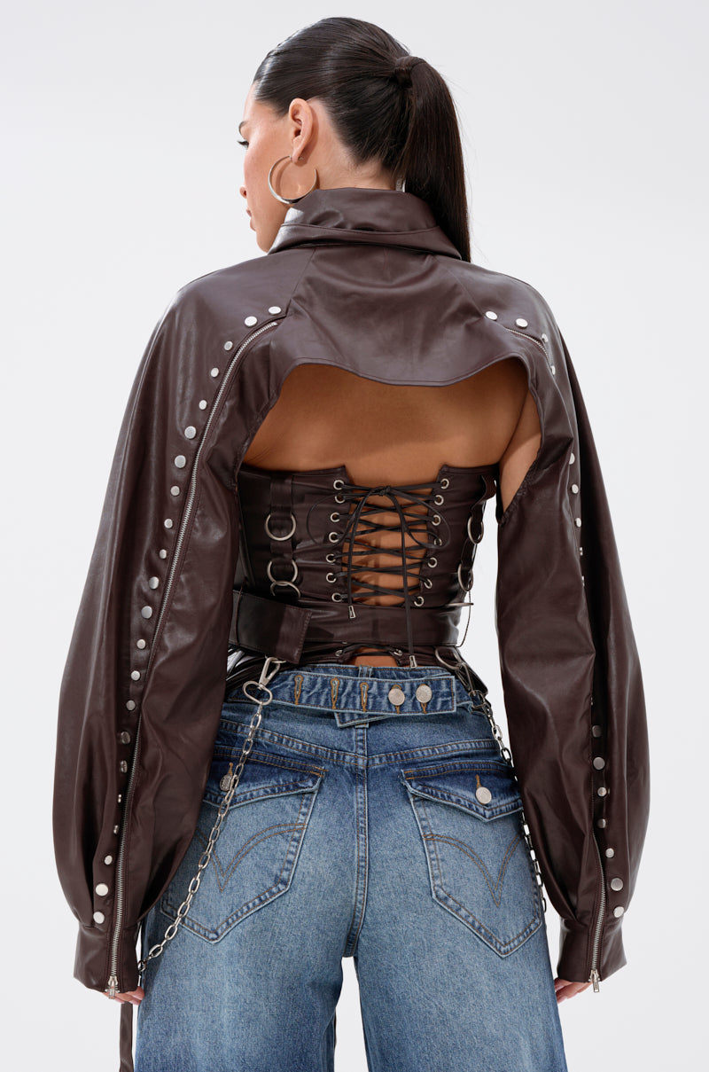 A woman with long dark hair in a ponytail wears the PERK UP EMBELLISHED BOLERO with silver studs and a cutout back, paired with high-waisted blue jeans featuring chain accents and lace-up corset-style lacing.
