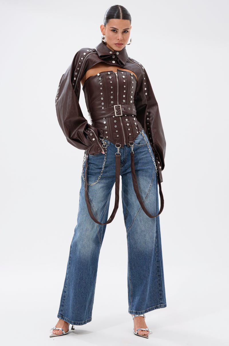 A woman stands confidently in the PERK UP EMBELLISHED BOLERO, a studded brown faux leather jacket with belted details, styled with wide-leg blue jeans, silver heels, and chain accessories against a plain white background.