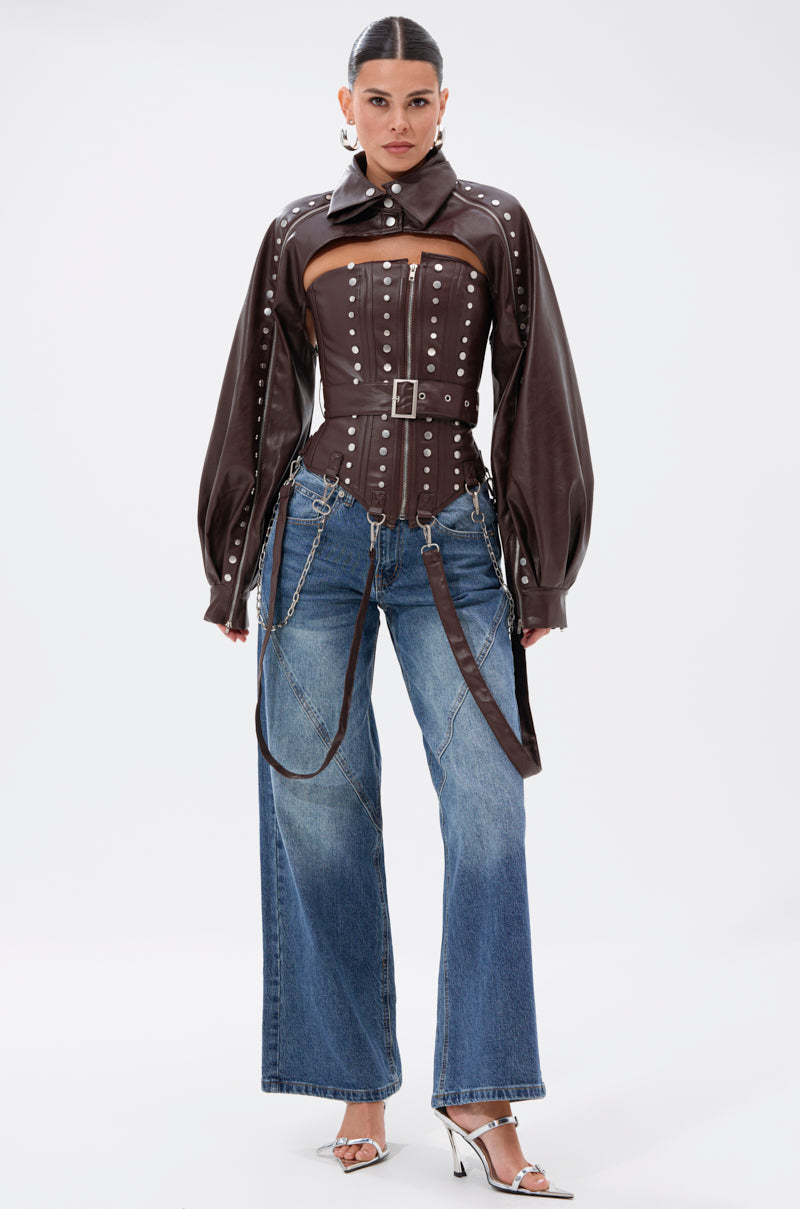 A woman faces forward, wearing the PERK UP EMBELLISHED BOLERO over a brown leather corset top with silver studs, wide-leg blue jeans, metallic heels, and chain accessories. Her hair is slicked back against a plain white background.