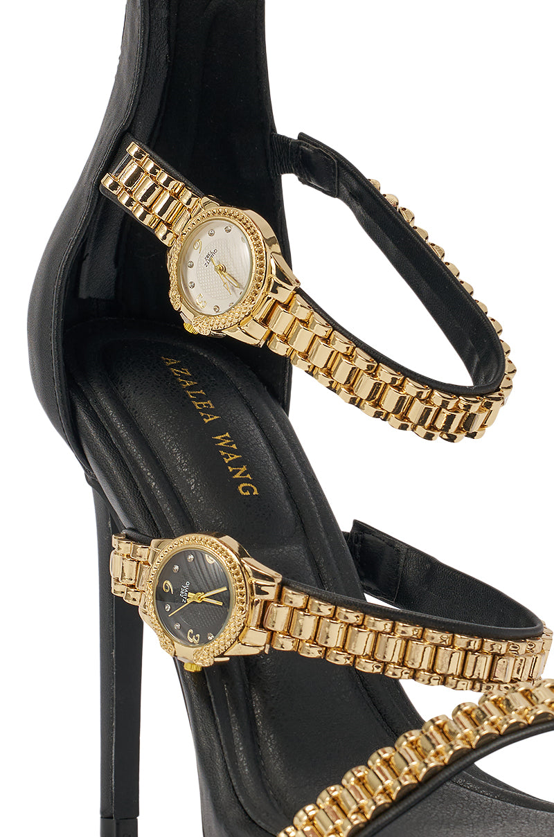 A close-up of the AZALEA WANG BEZEL WATCH STRAP NOVELTY STILETTO SANDAL in black, featuring two gold watch-link straps, each set with a small watch face—one white and one black. 6