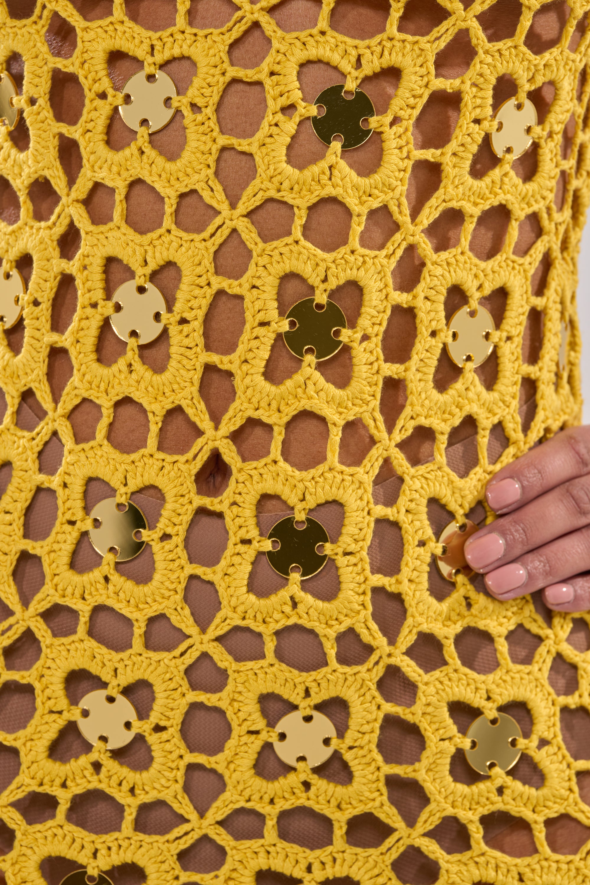 A close-up of a hand resting on the GLOW UP SEQUIN CROCHET MINI DRESS, featuring metallic gold flower-shaped sequins, intricate open-weave crochet, and nude-colored nails. 8