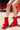 Wearing the AZALEA WANG BHARTI RED SNAP DETAIL BOOTIE, a person with white socks with yellow stripes sits on a red picnic bench outdoors; only their legs and feet are visible.