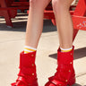 Wearing the AZALEA WANG BHARTI RED SNAP DETAIL BOOTIE, a person with white socks with yellow stripes sits on a red picnic bench outdoors; only their legs and feet are visible.