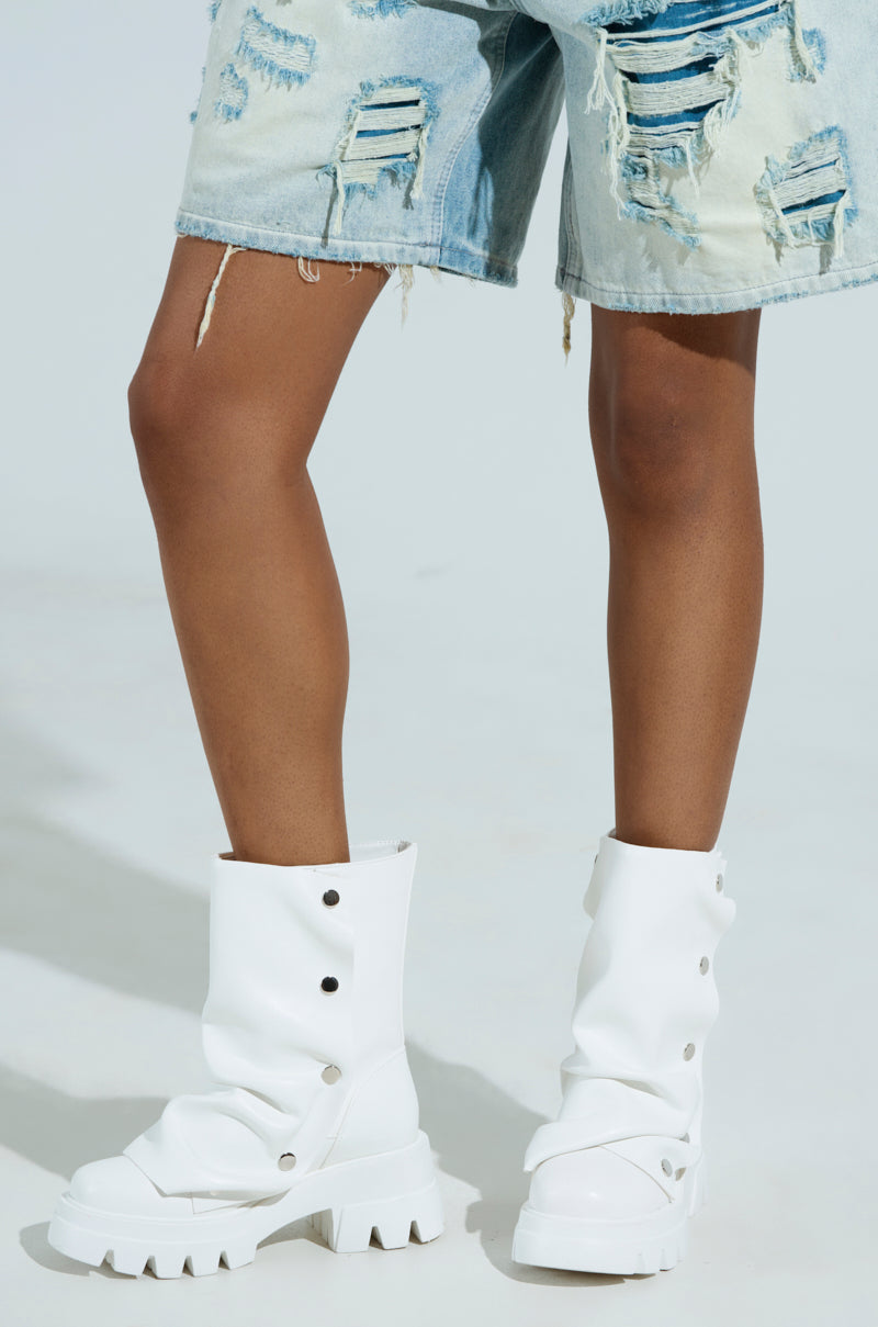 A person stands wearing distressed denim shorts paired with the AZALEA WANG BHARTI WHITE SNAP BOOTIE—tall white vegan leather boots with chunky heels and snap details—offering a stylish edge. The loose ankle fit contrasts against a plain light background.