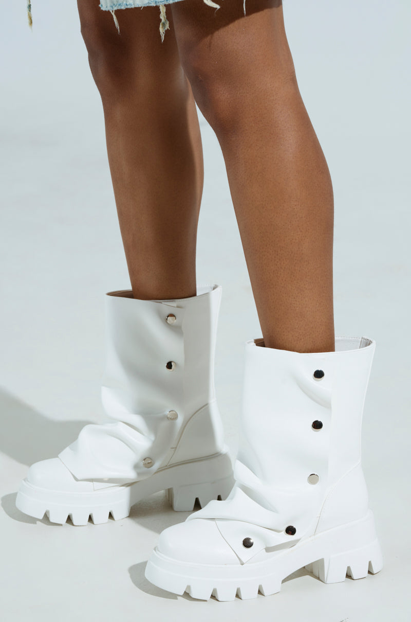 A person stands on a light background wearing AZALEA WANG BHARTI WHITE SNAP BOOTIE, featuring oversized white vegan leather with chunky heels and multiple snap buttons. Their legs are visible from the knees down, and the booties have a slightly slouching effect.